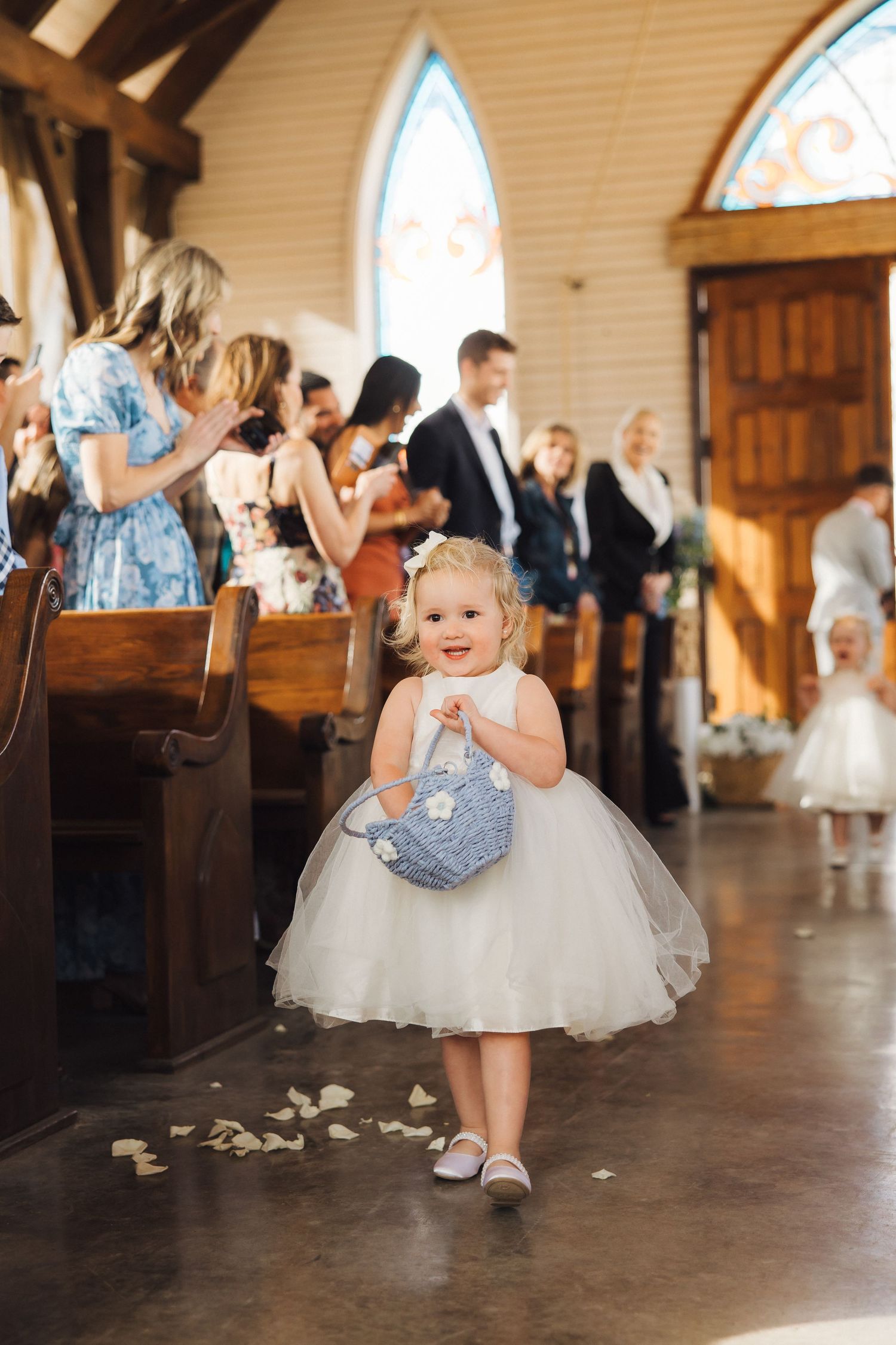 A young flower girl in a white tulle dress walks down the aisle during a church wedding ceremony.