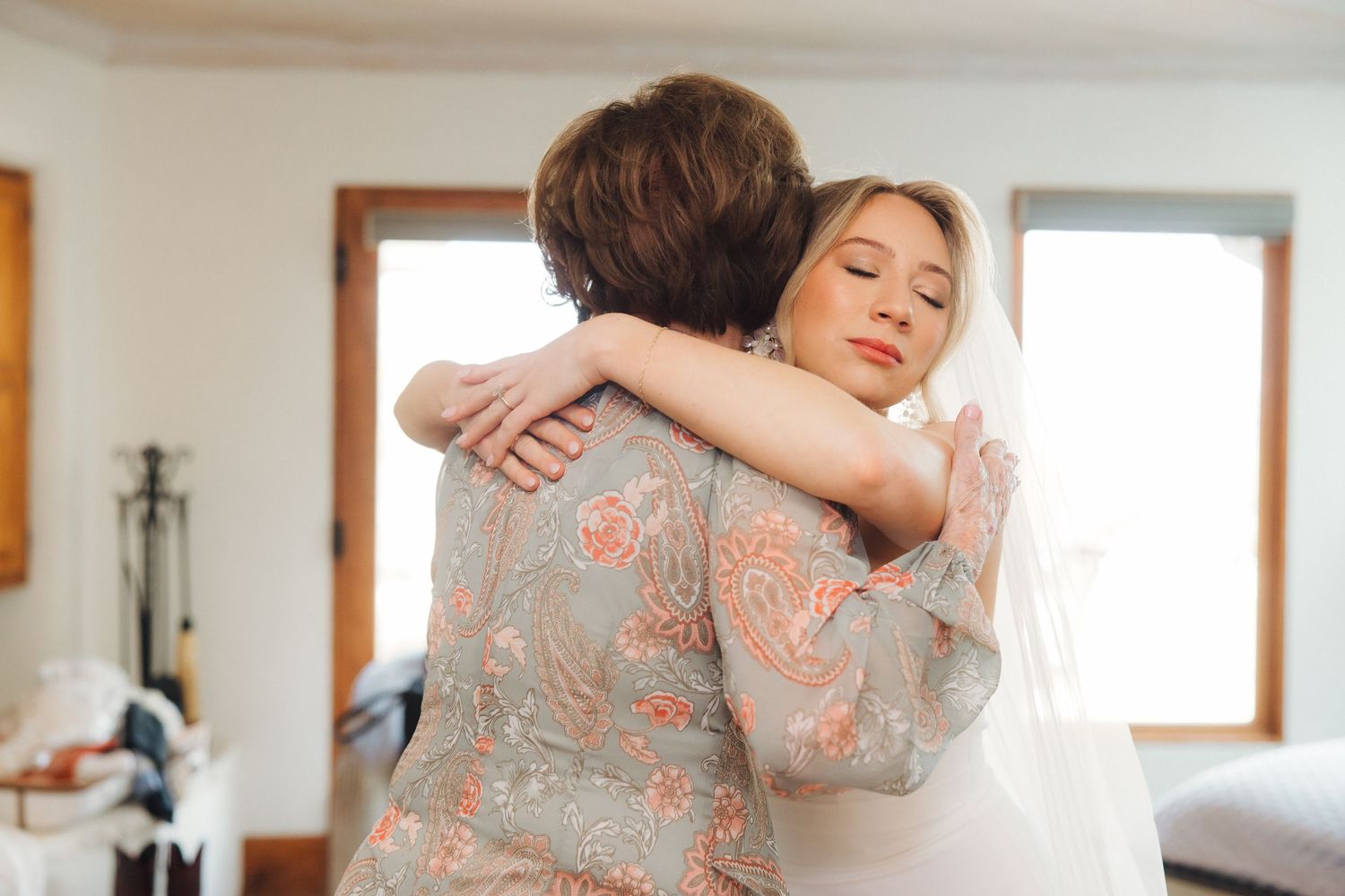 Two people share an emotional embrace in a sunlit room while one wears a patterned dress with floral details.