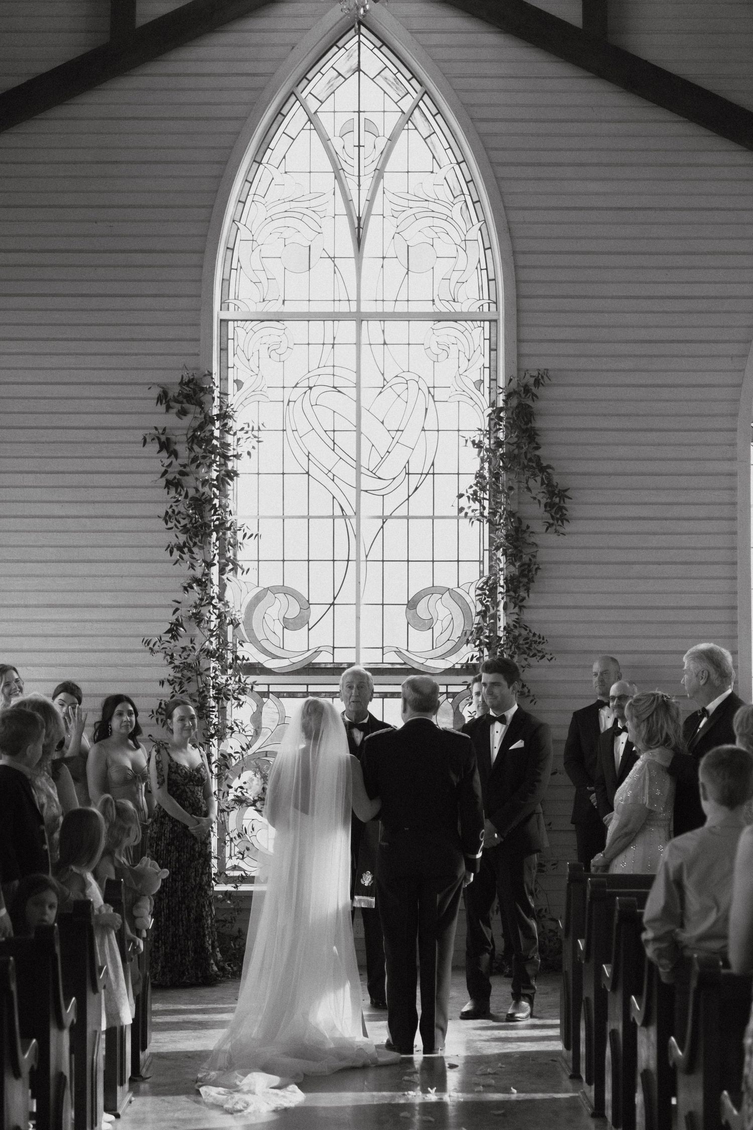 Black and white panorama of a wedding ceremony inside a chapel with a beautiful arched stained glass window.