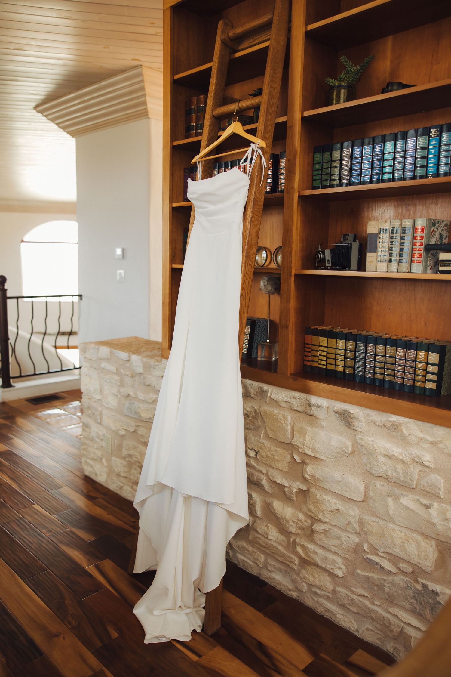Elegant white wedding dress hanging on wooden hanger against stone wall and bookshelf in rustic interior.