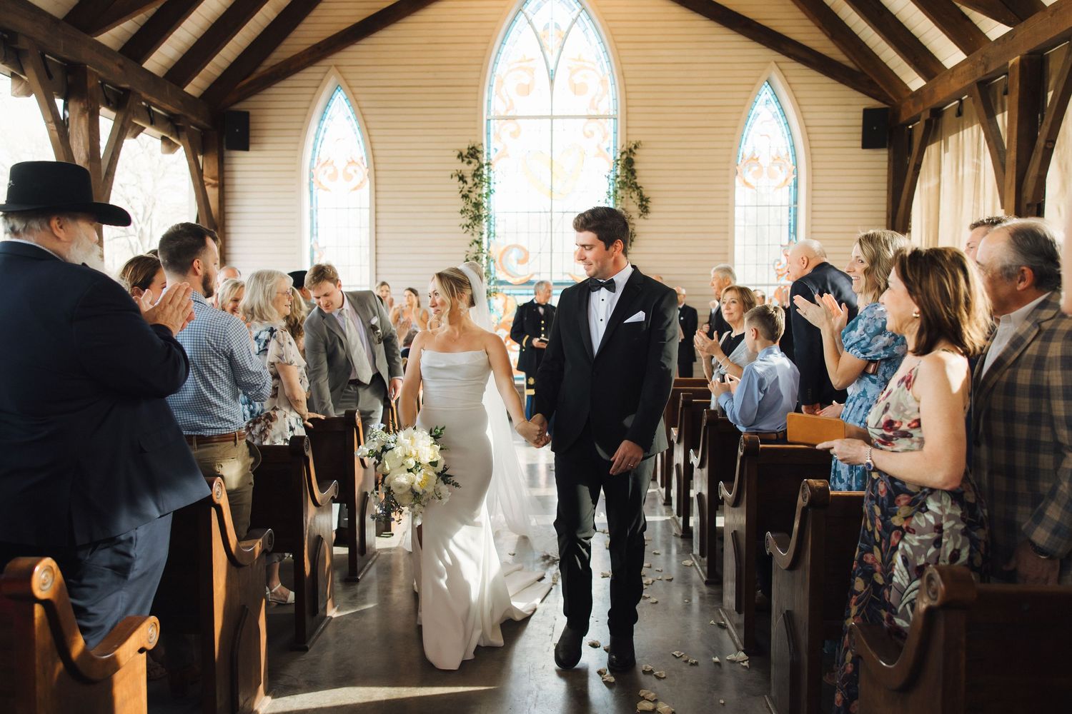 Wedding ceremony in a charming chapel with gothic arched windows as guests celebrate the newlyweds walking down the aisle.