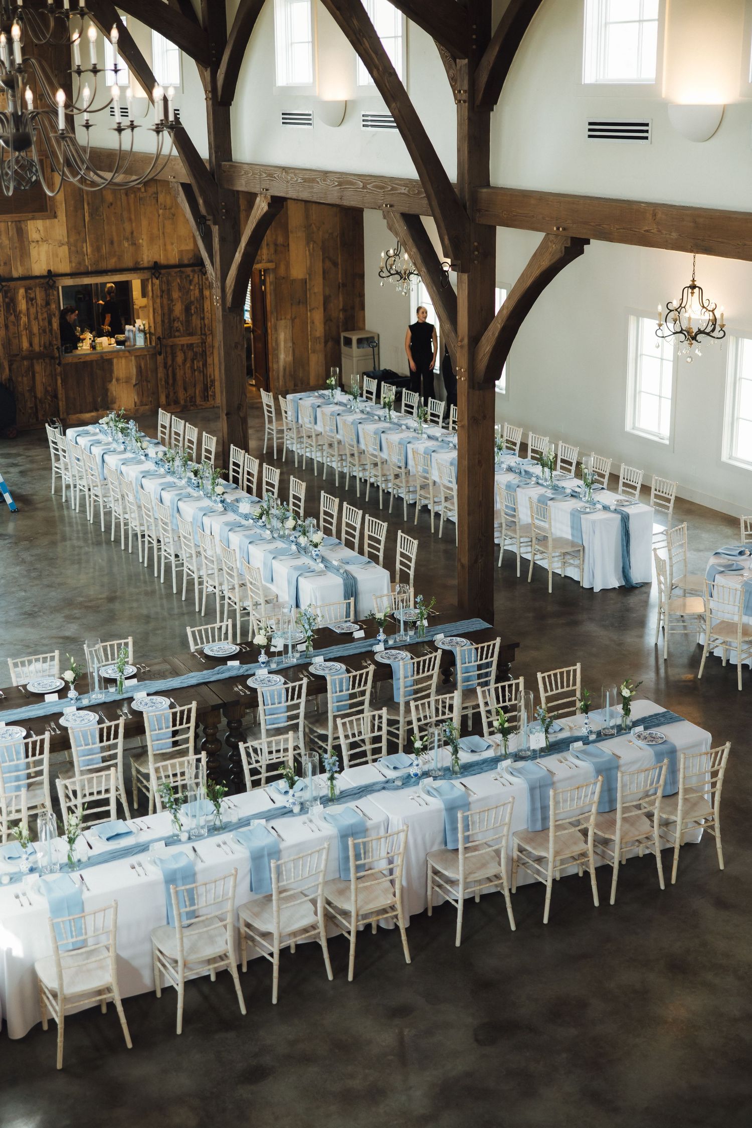 Long banquet tables with white linens and chiavari chairs are arranged in rows inside a rustic barn reception venue.
