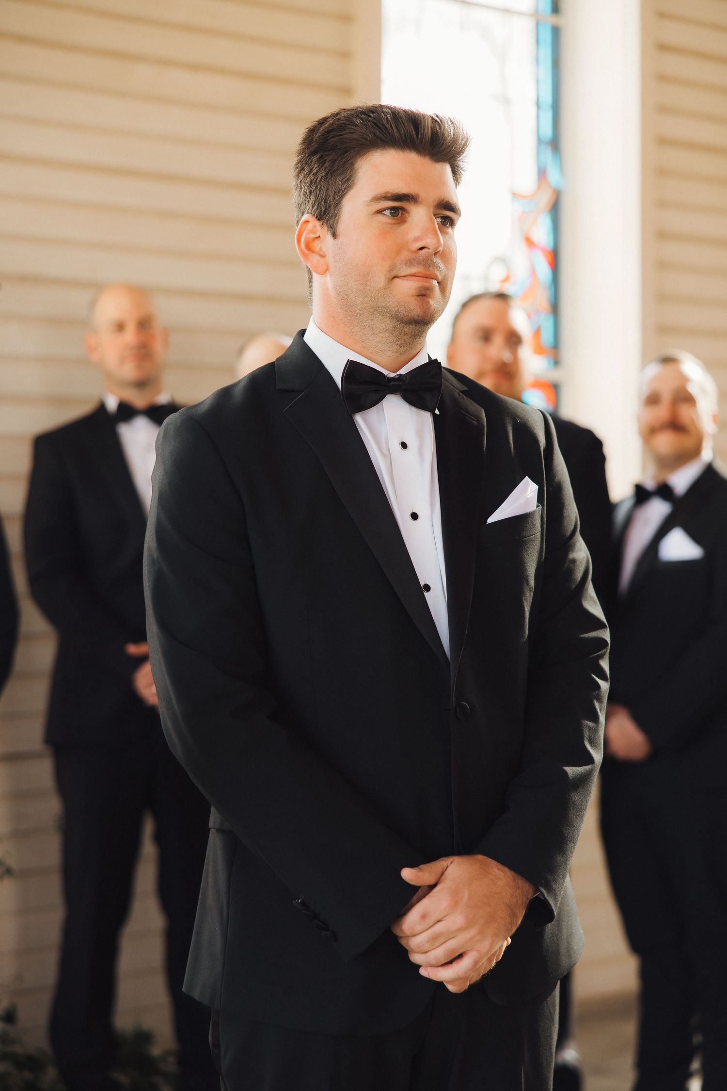 A groom in a classic black tuxedo stands waiting in front of stained glass windows.
