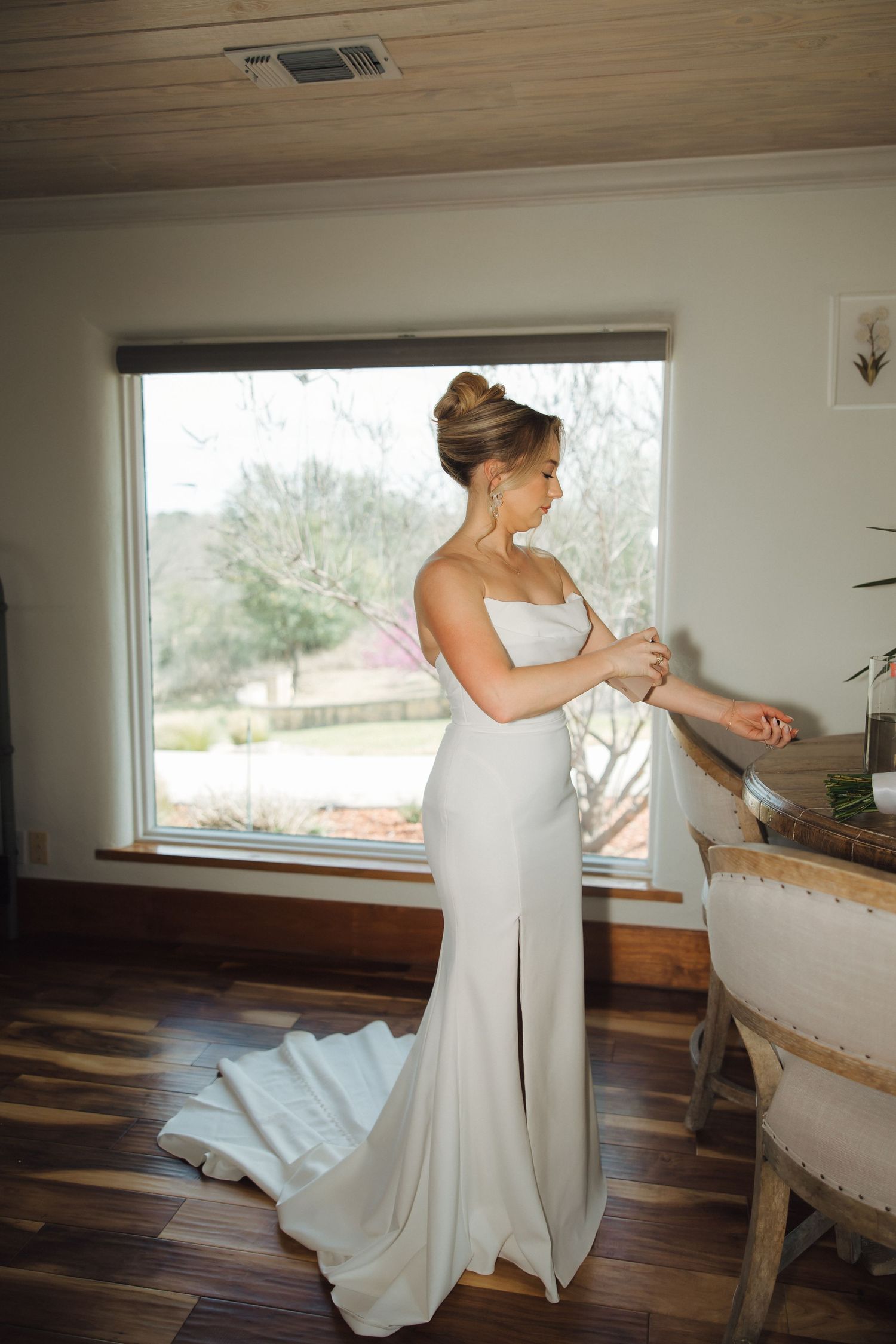 Elegant bride in fitted white gown adjusting jewelry by window in natural light.