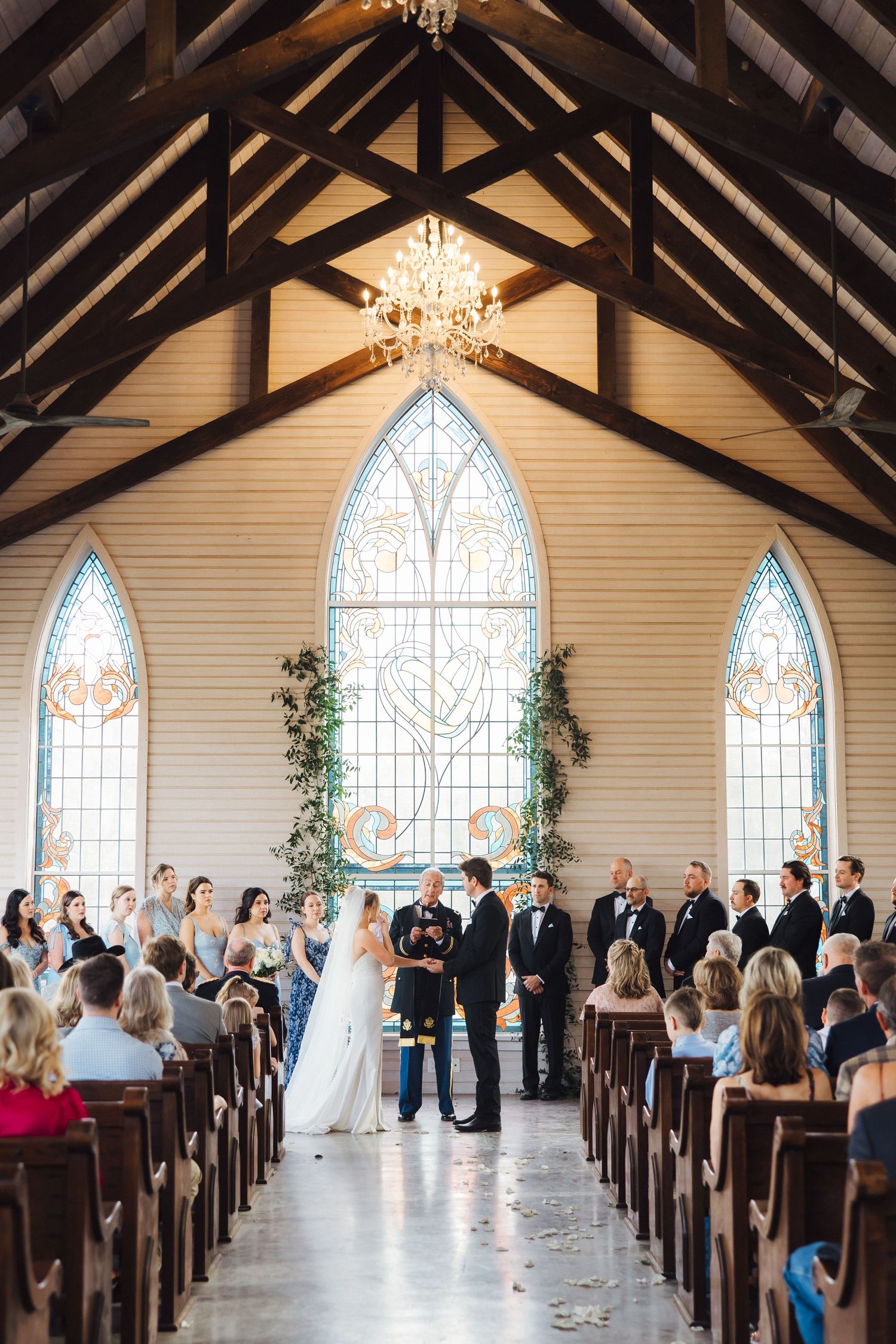 A wedding ceremony view from the back of a chapel showing vaulted ceilings and stunning stained glass windows.