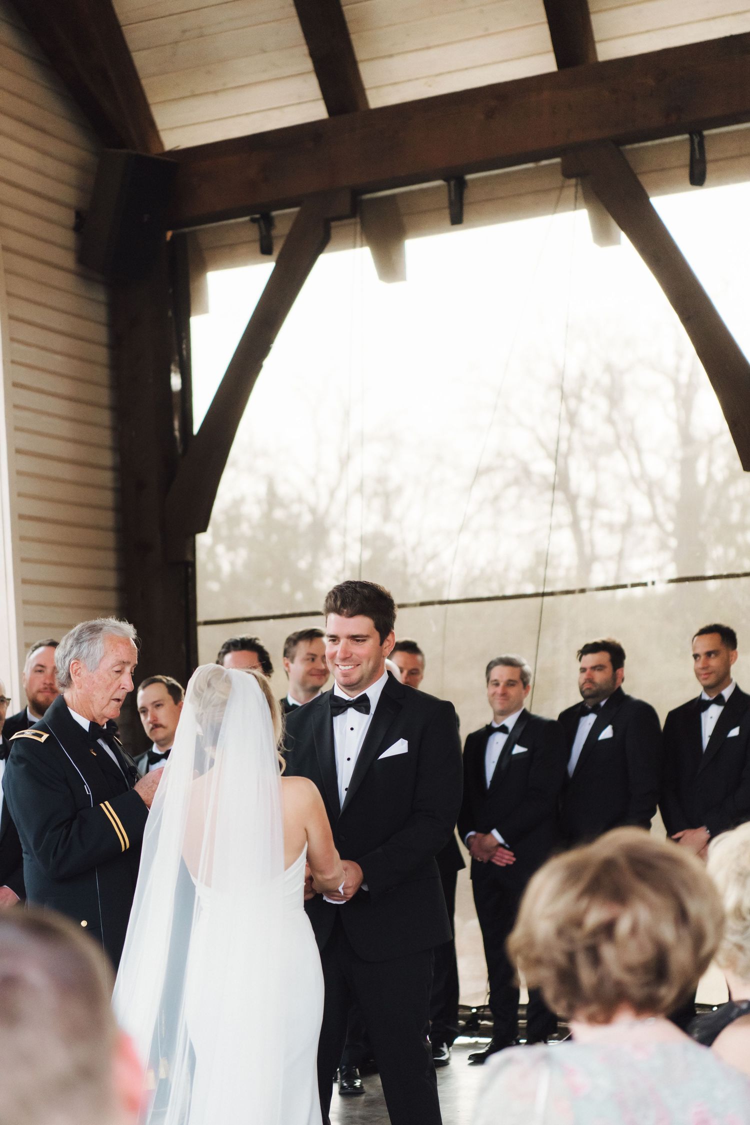 Wedding party in formal attire stands in a line under a covered outdoor pavilion.