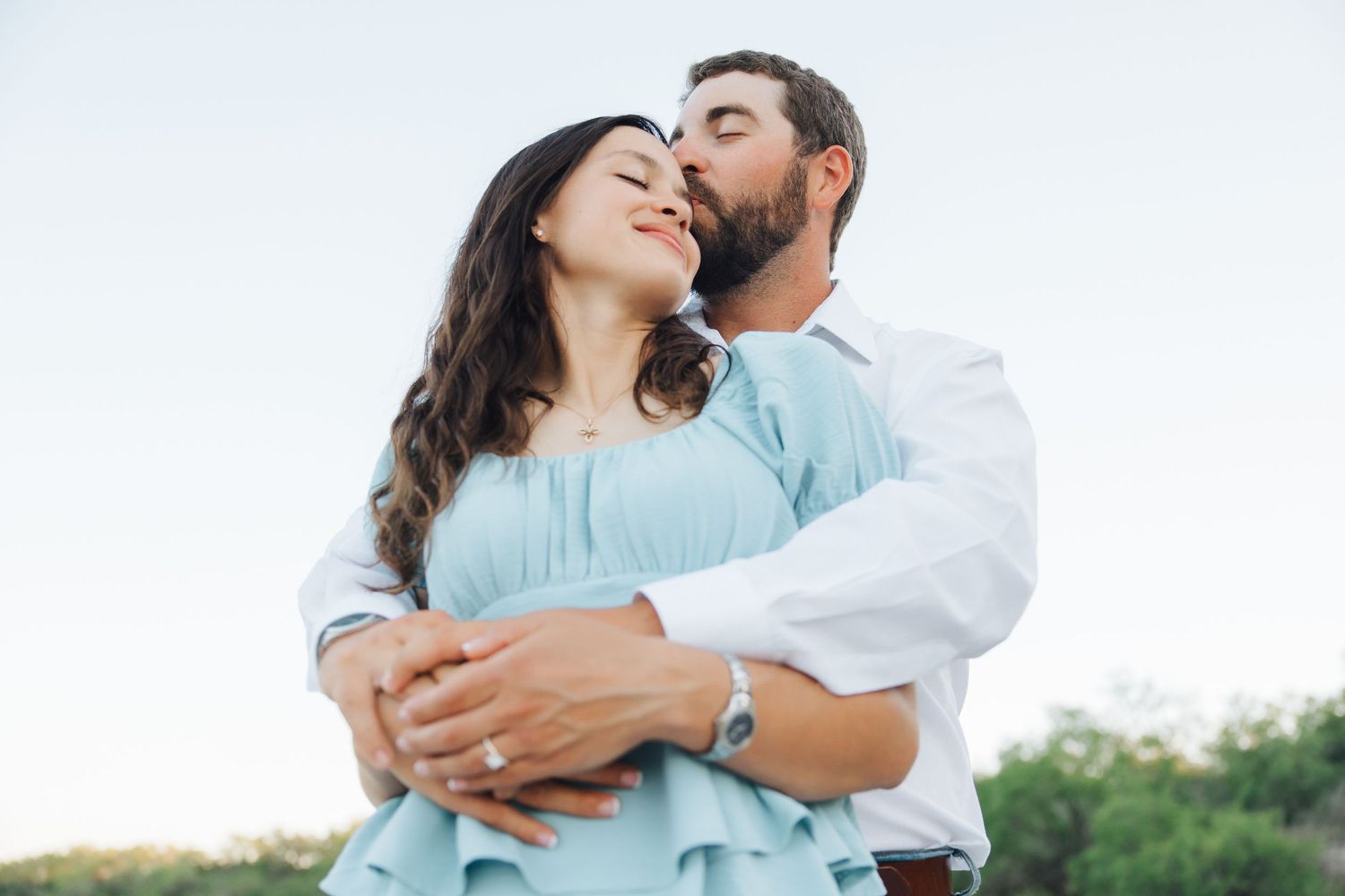 A couple in white and light blue clothing embrace tenderly during a romantic outdoor photo session.