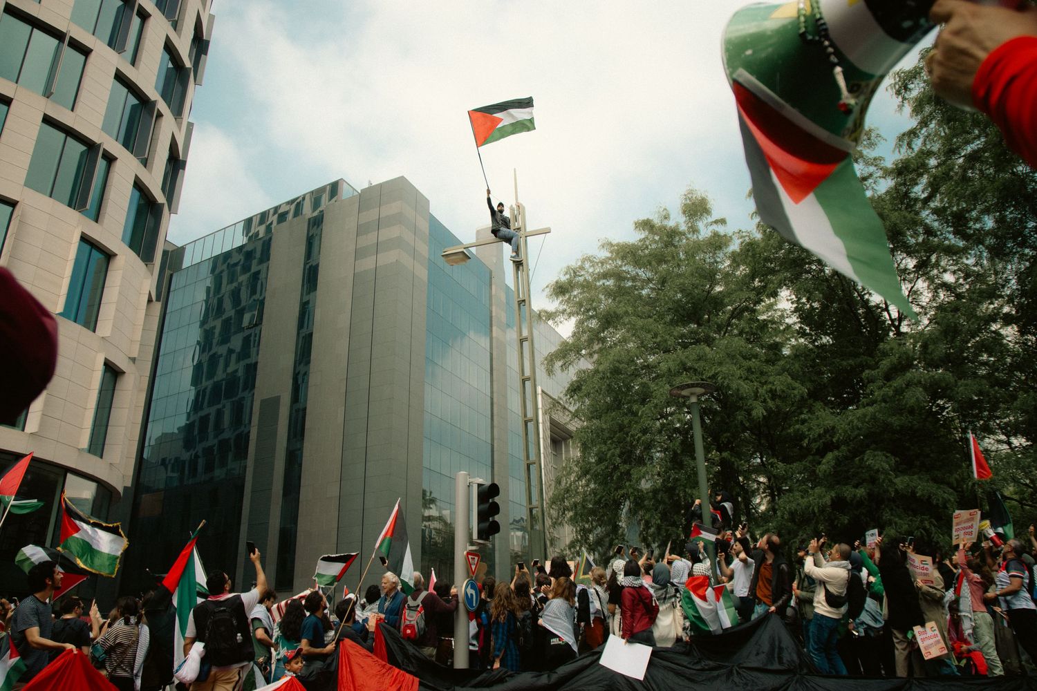 A demonstration for Palestine in Brussels. A man sitting on a tall pole, waving a Palestinian flag while the crowd cheers.