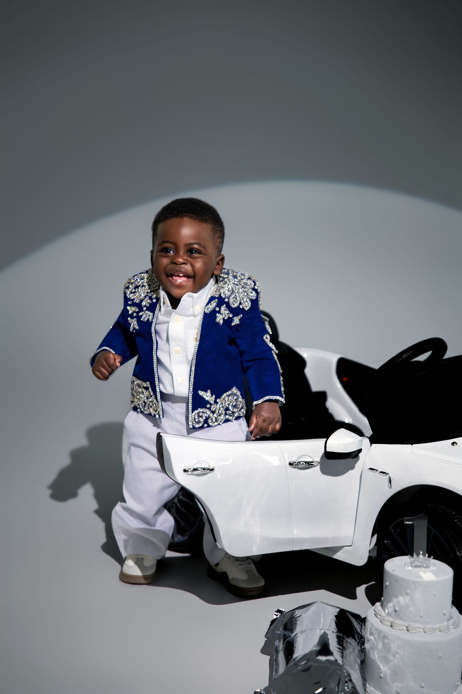 Smiling toddler in blue embroidered mariachi suit stands beside white toy car in studio setting.