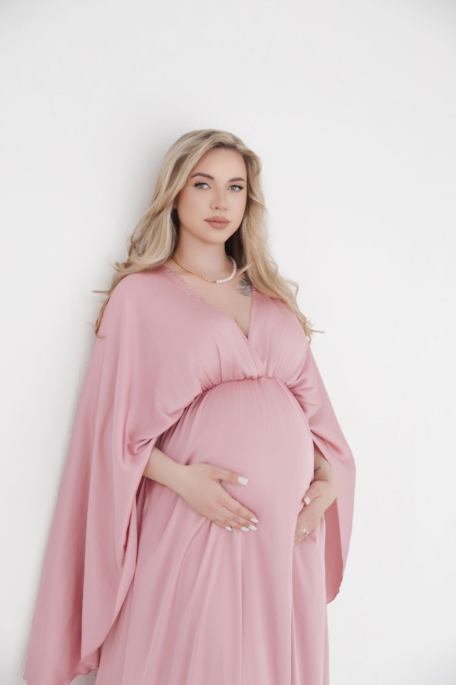 Elegant pregnant woman in pink flowing dress cradling baby bump against white background.