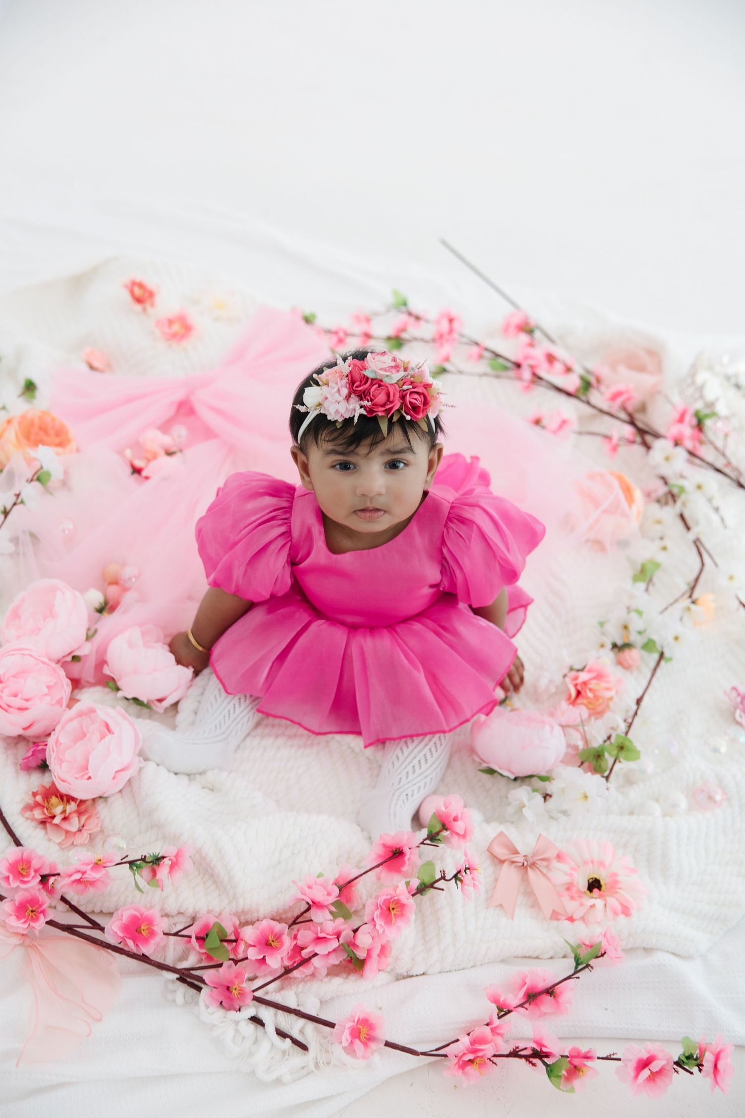 A baby girl in a pink tutu dress and floral headband sits surrounded by pink flowers on a white backdrop.