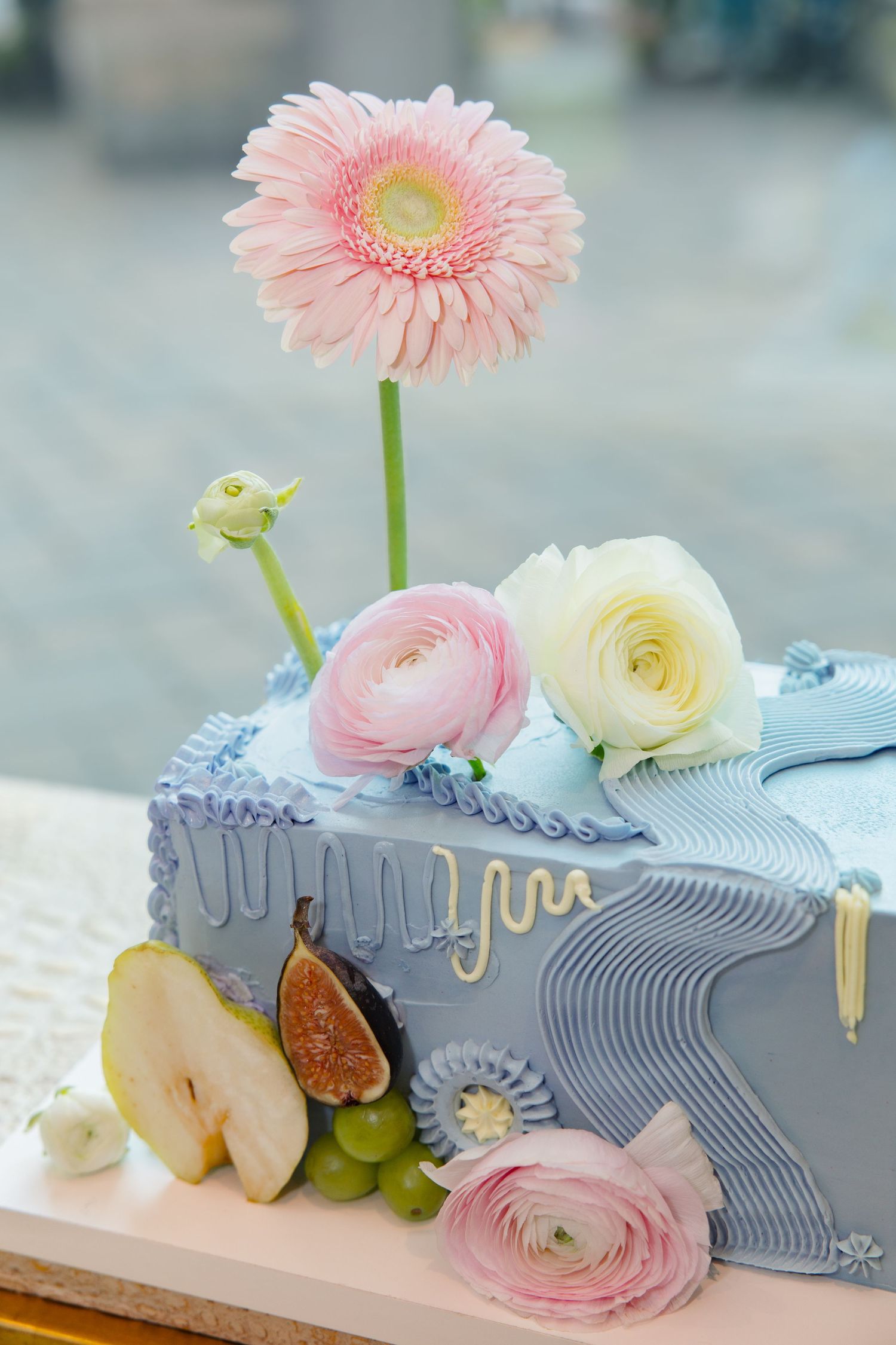 Elegant blue fondant cake decorated with pink gerbera daisy, ranunculus, roses, fresh figs, pear slices, and green grapes.