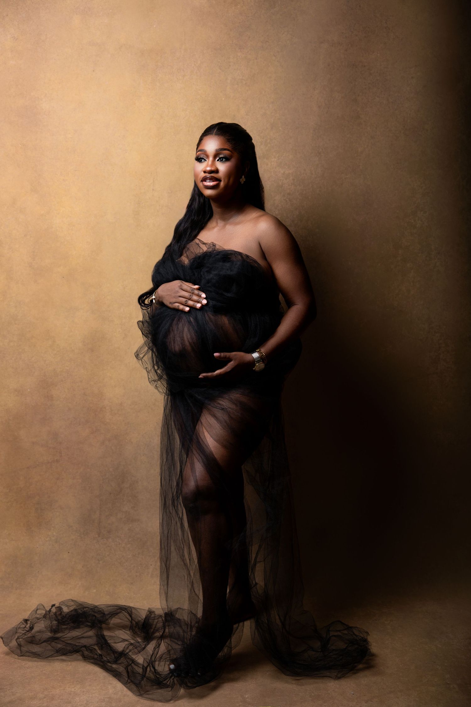 Pregnant woman in black tulle gown cradling baby bump during elegant maternity photo session on golden background.