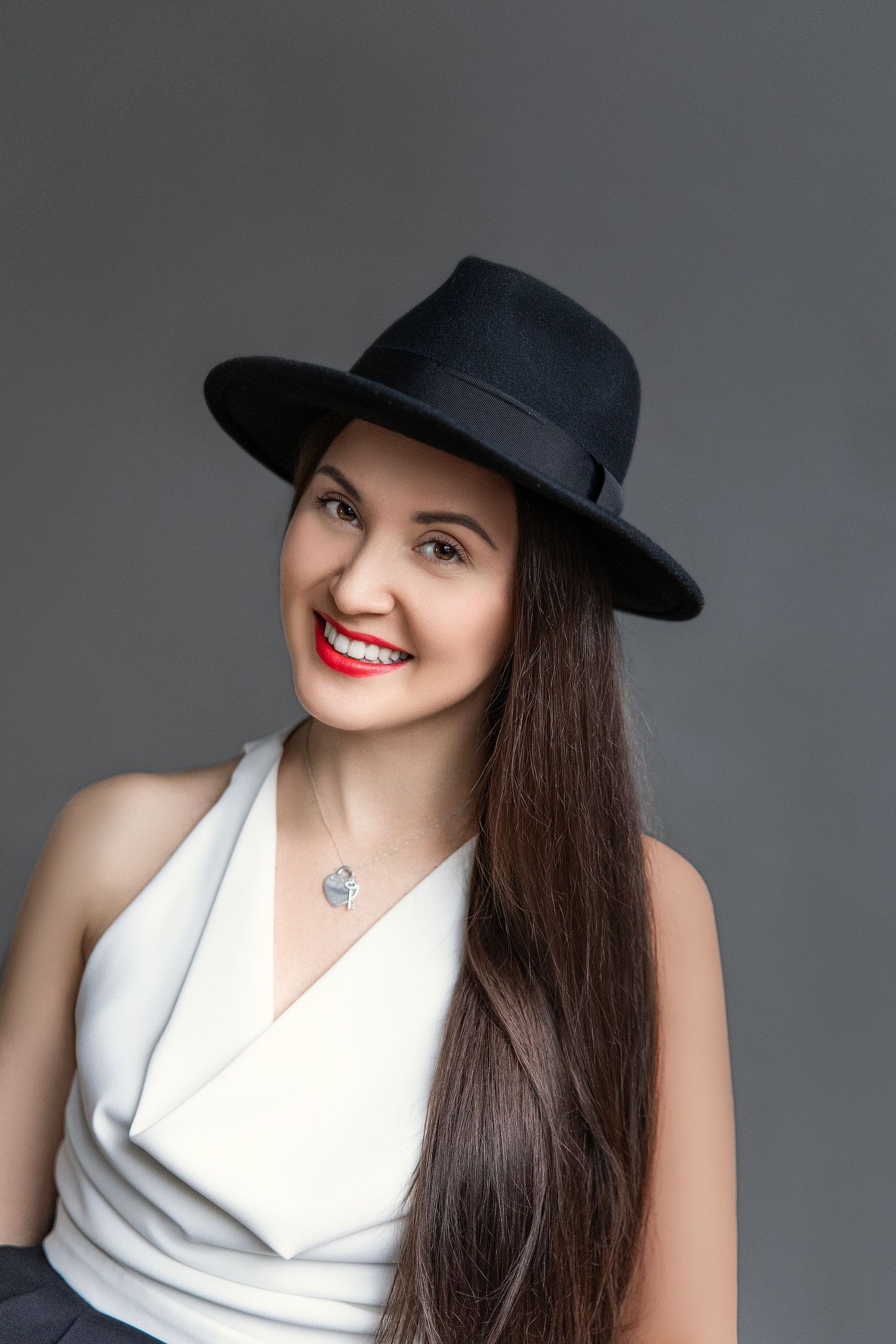 Elegant woman with long dark hair and red lips smiling in black fedora hat and white dress top.