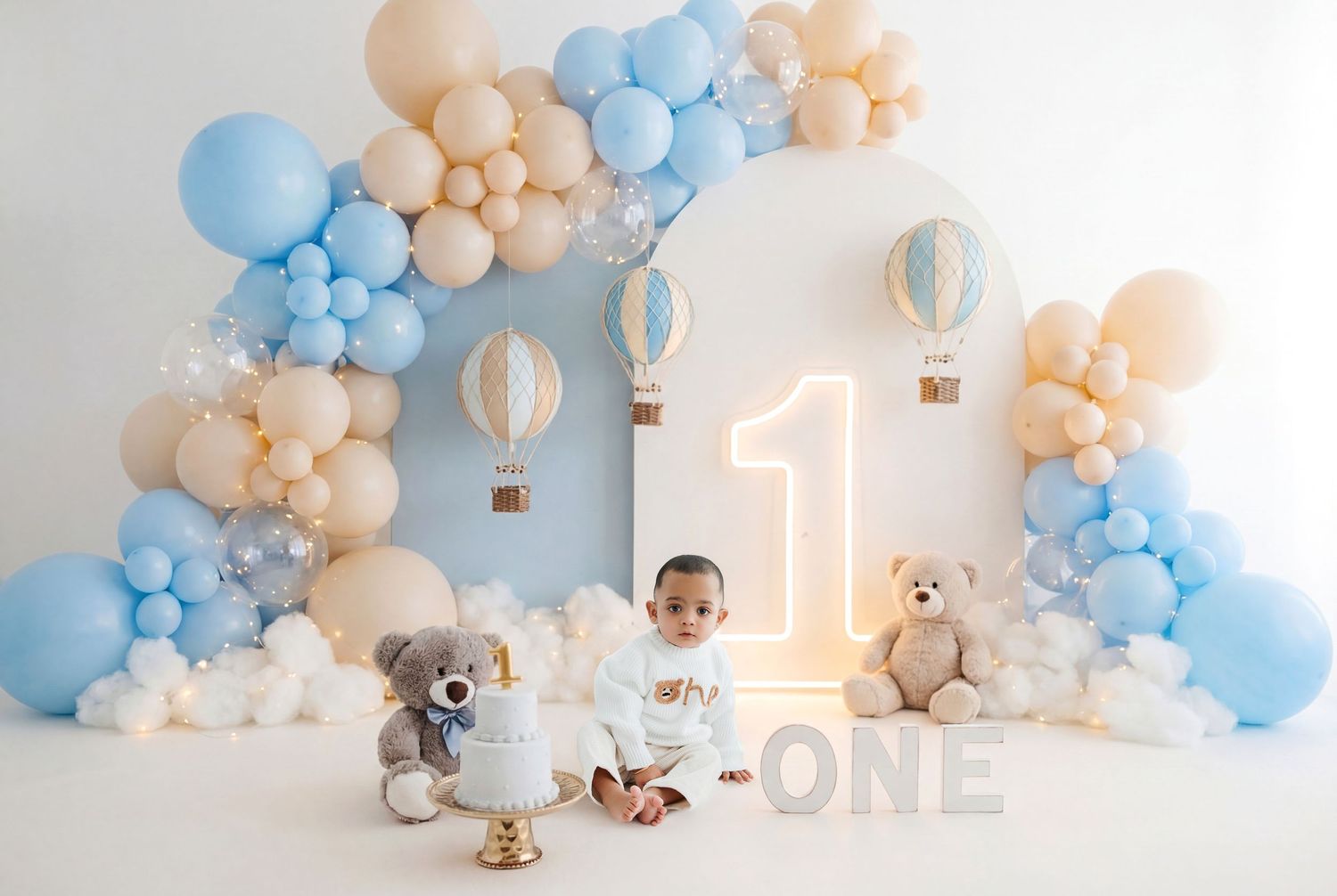 Baby's first birthday setup with blue and beige balloons, teddy bears, neon number one, and hot air balloon decorations.