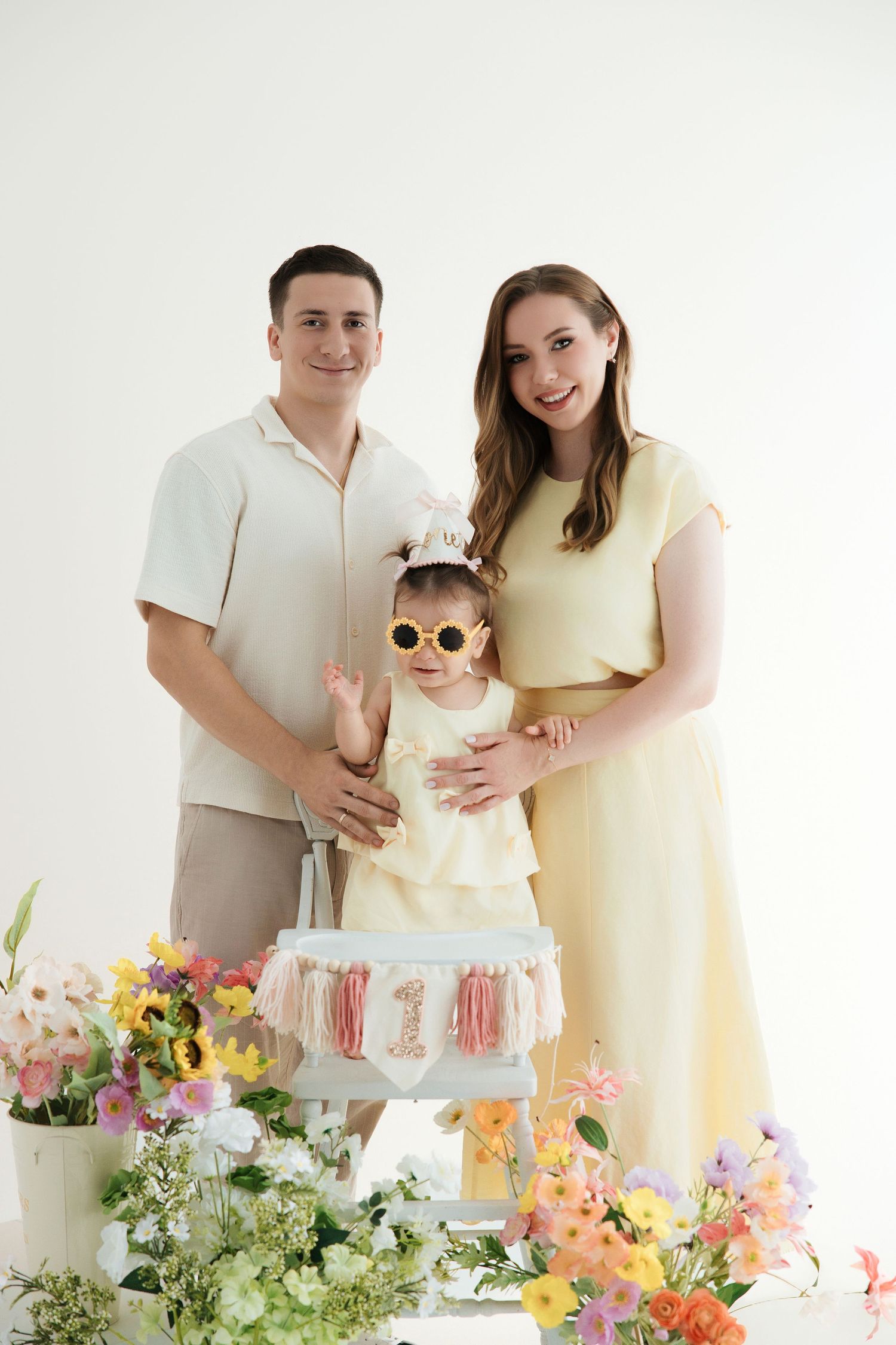 A family of three poses with colorful flowers and a first birthday cake, baby girl wearing sunglasses and crown.