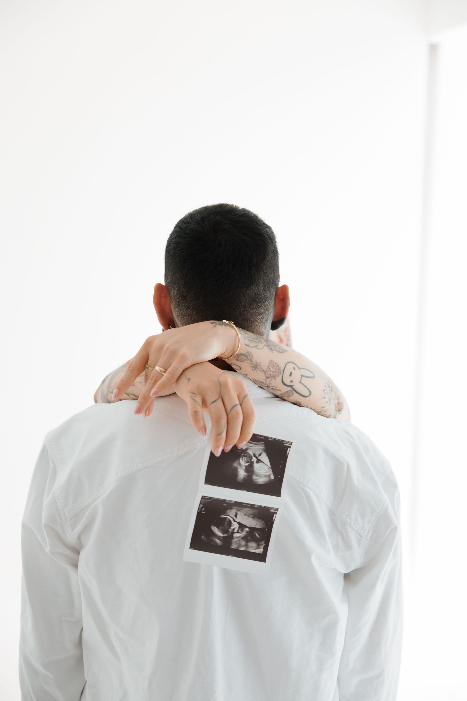 A man with neck tattoos holds ultrasound photos on his back while a woman with rings wraps her arms around him.