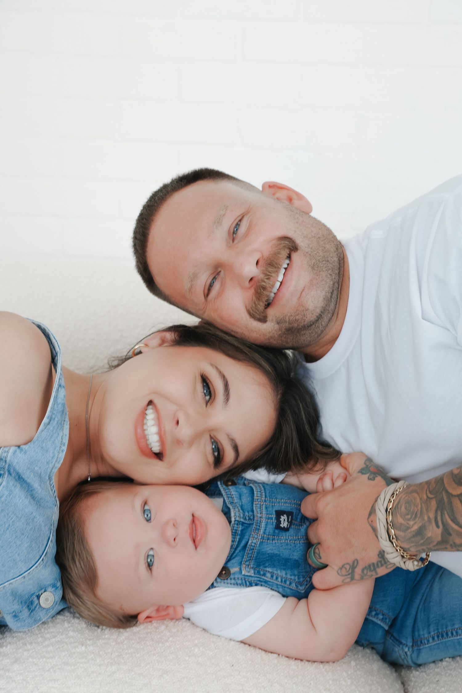 A happy young family with tattooed father, smiling mother, and baby in denim outfits posing together on a white background.