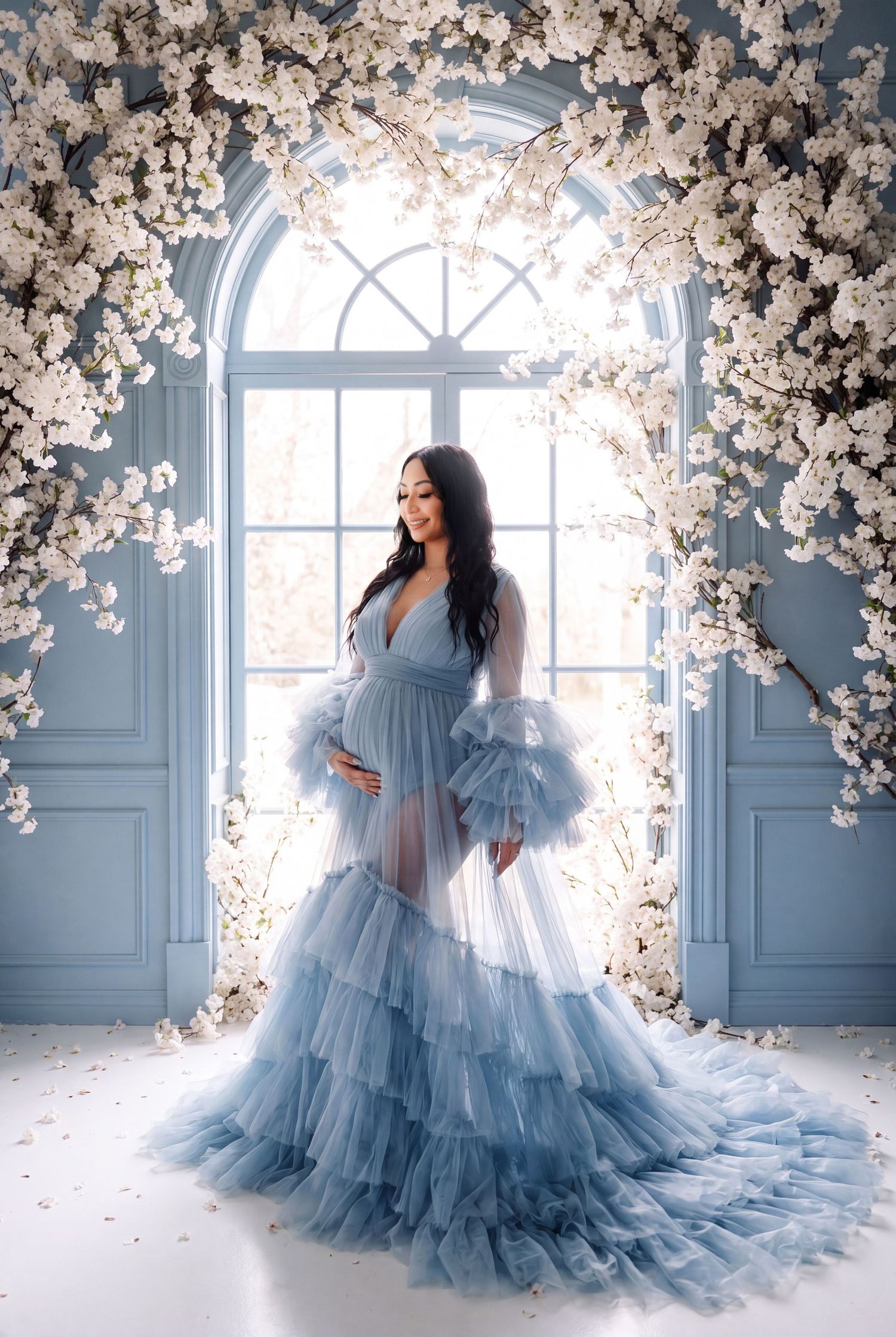 Pregnant woman in blue tulle gown poses under white floral arch in elegant blue studio setting.