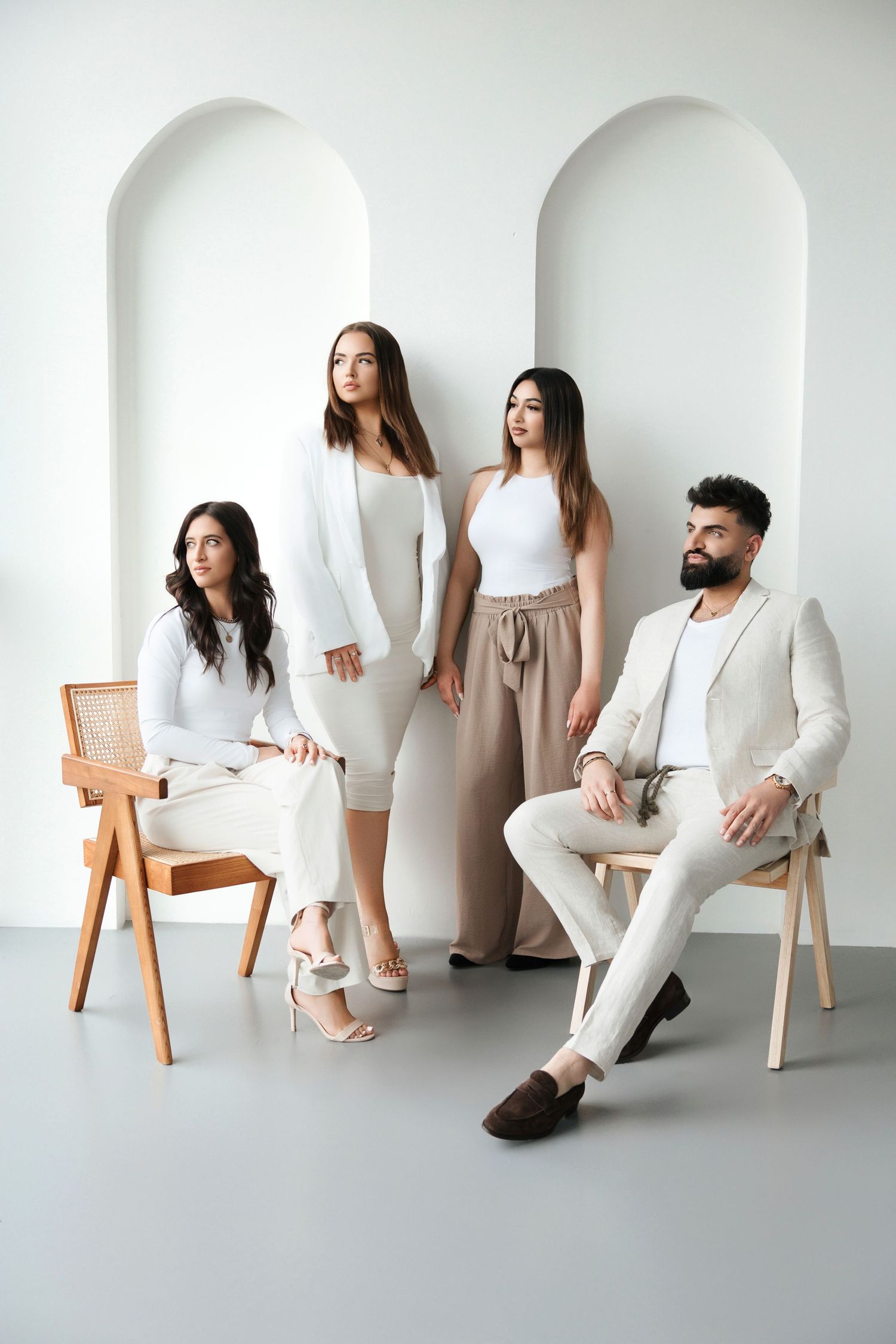 Four fashionable people in white and beige outfits posing with wooden chairs against arched white studio background.