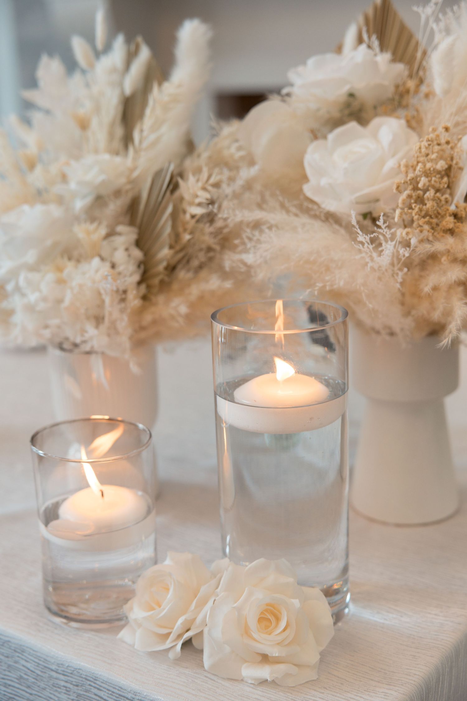Elegant wedding centerpiece with floating candles, white roses, and dried pampas grass floral arrangements on a white table.