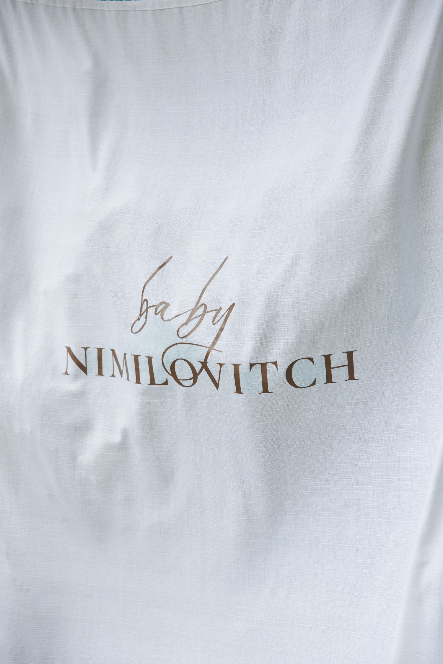 White fabric with gold cursive baby Nimilovitch brand logo printed in the center.