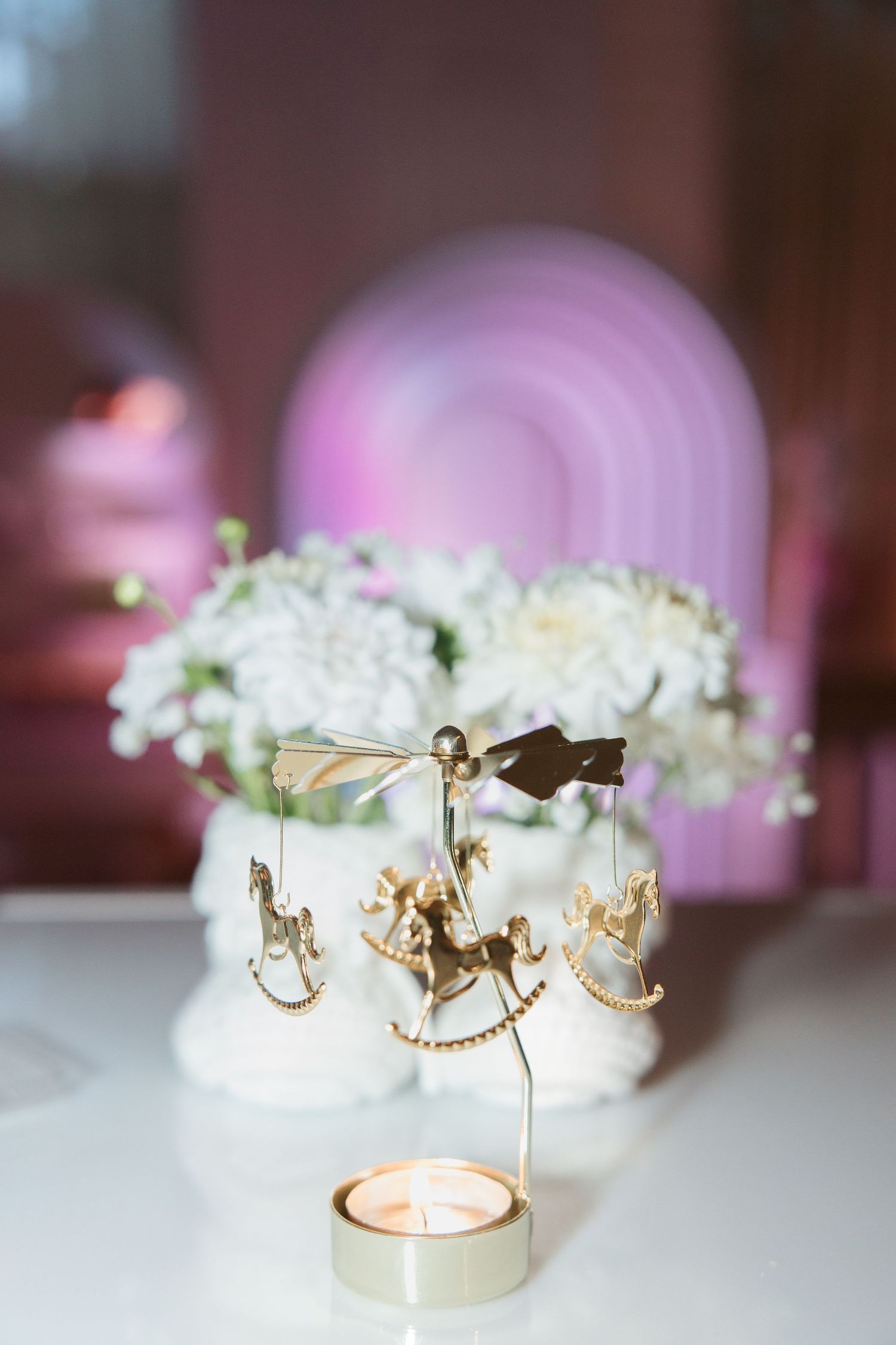 Gold carousel candle holder with rocking horses beside white chrysanthemums in soft pink bokeh background.