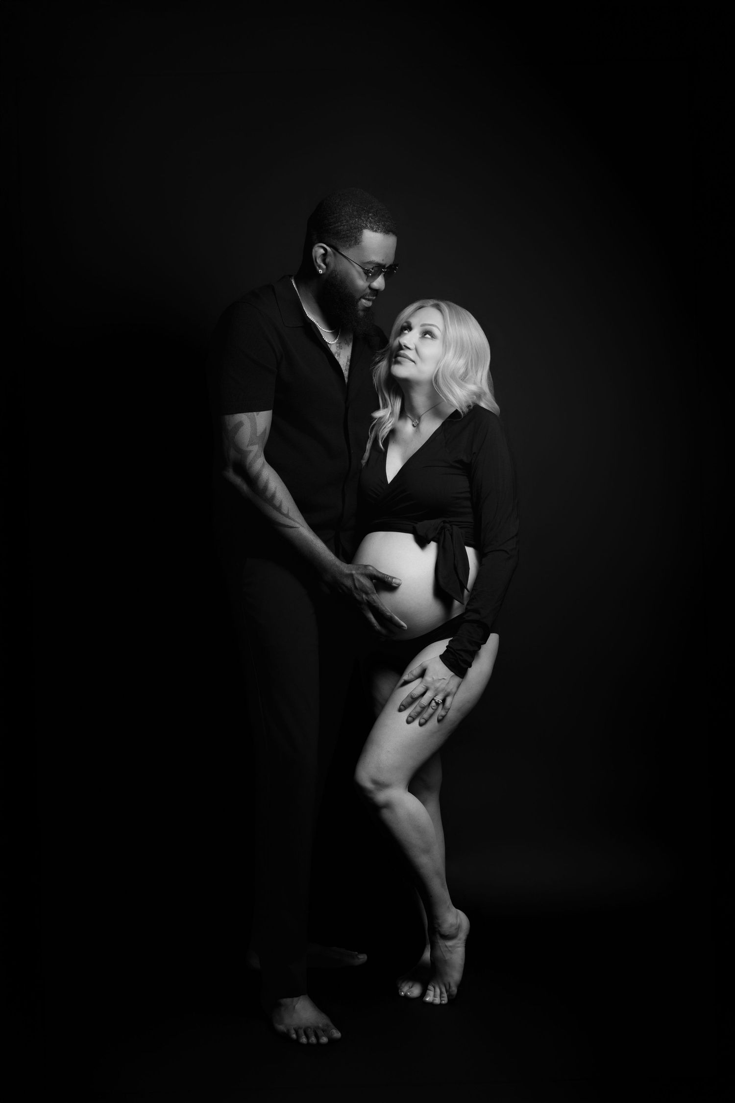 Black and white maternity photo of a couple, man embracing pregnant woman from behind in a studio.