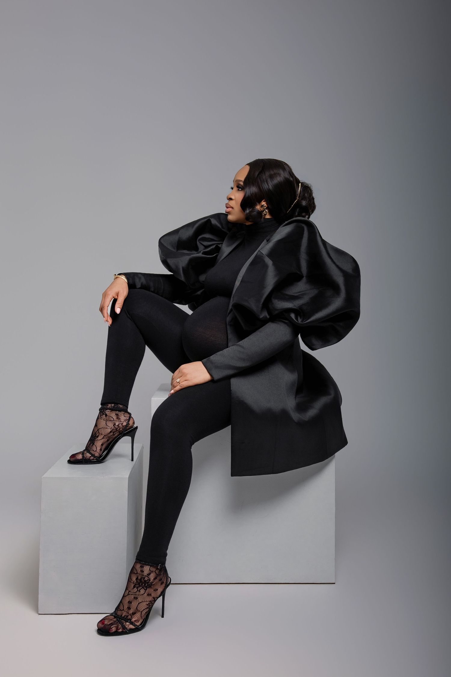 Woman in black satin sculptural jacket, leggings, and lace heels sits on white cube against gray background.