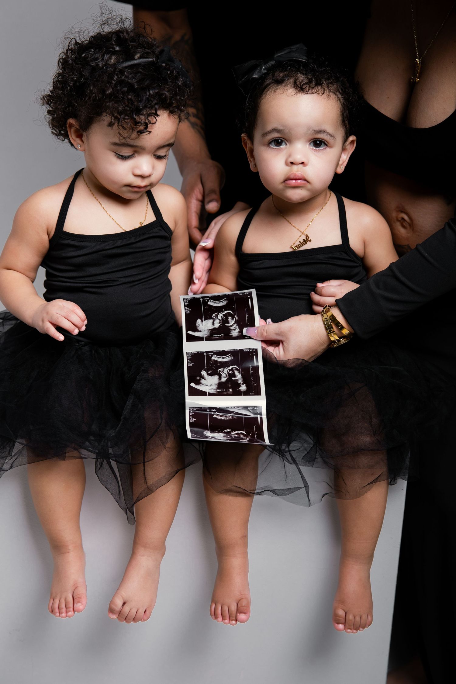 Two toddler girls in black tutu dresses hold ultrasound photos, announcing a new sibling pregnancy.