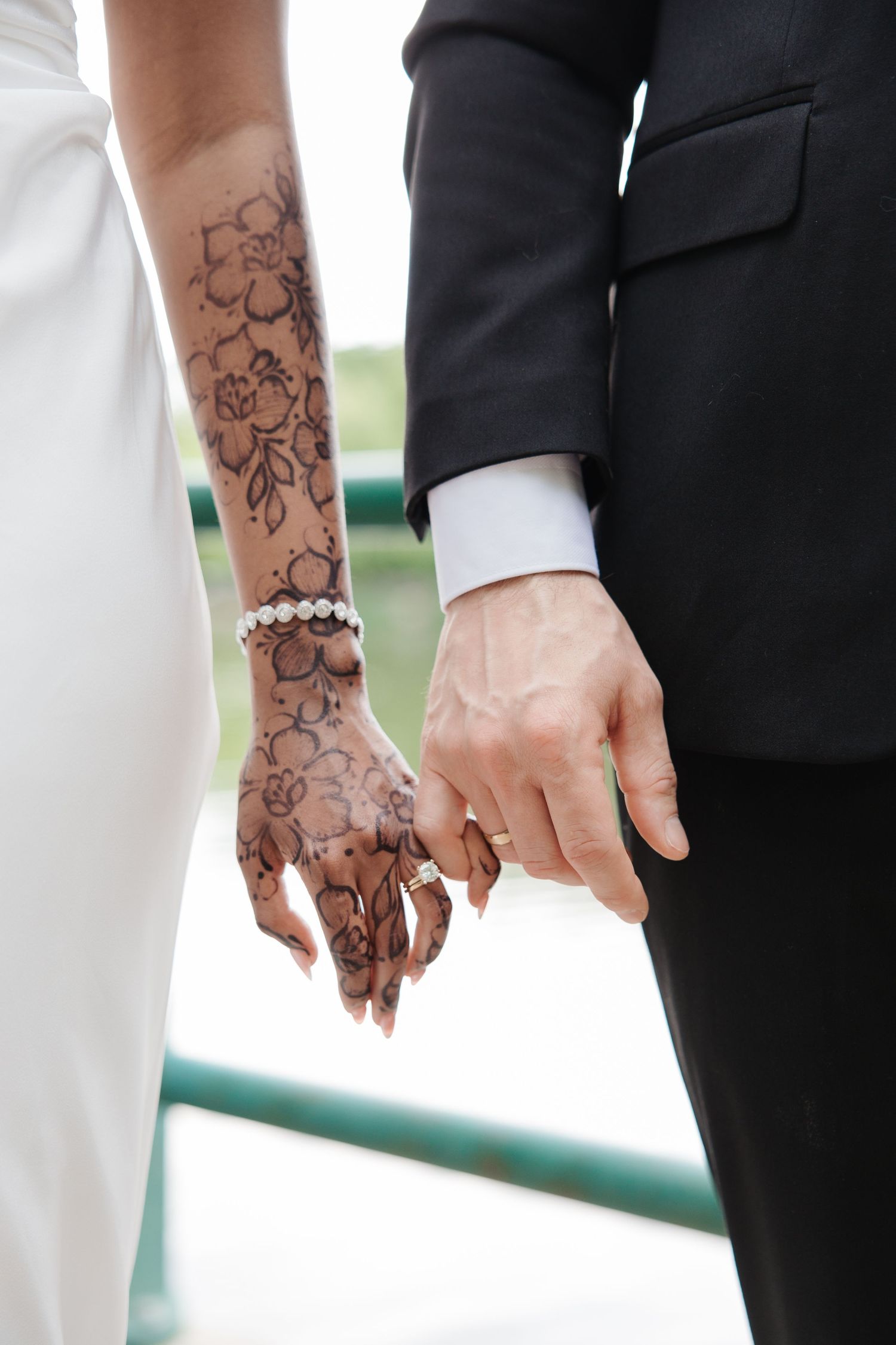 Bride with floral henna tattoo and pearl bracelet holding hands with groom in black suit at wedding ceremony.
