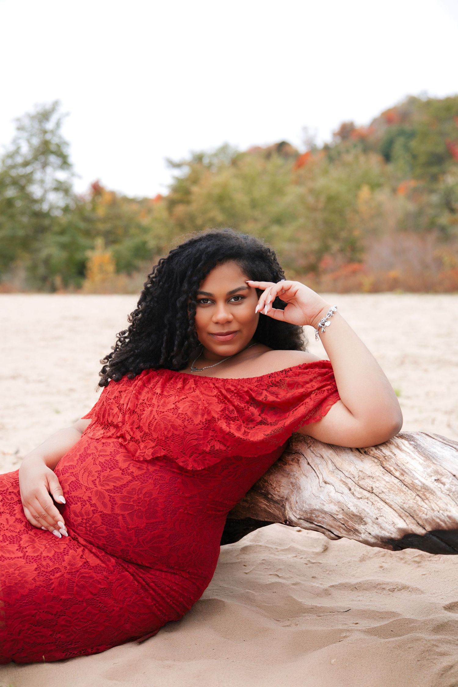 Woman in red off-shoulder dress poses on sandy beach leaning against driftwood with autumn foliage backdrop.