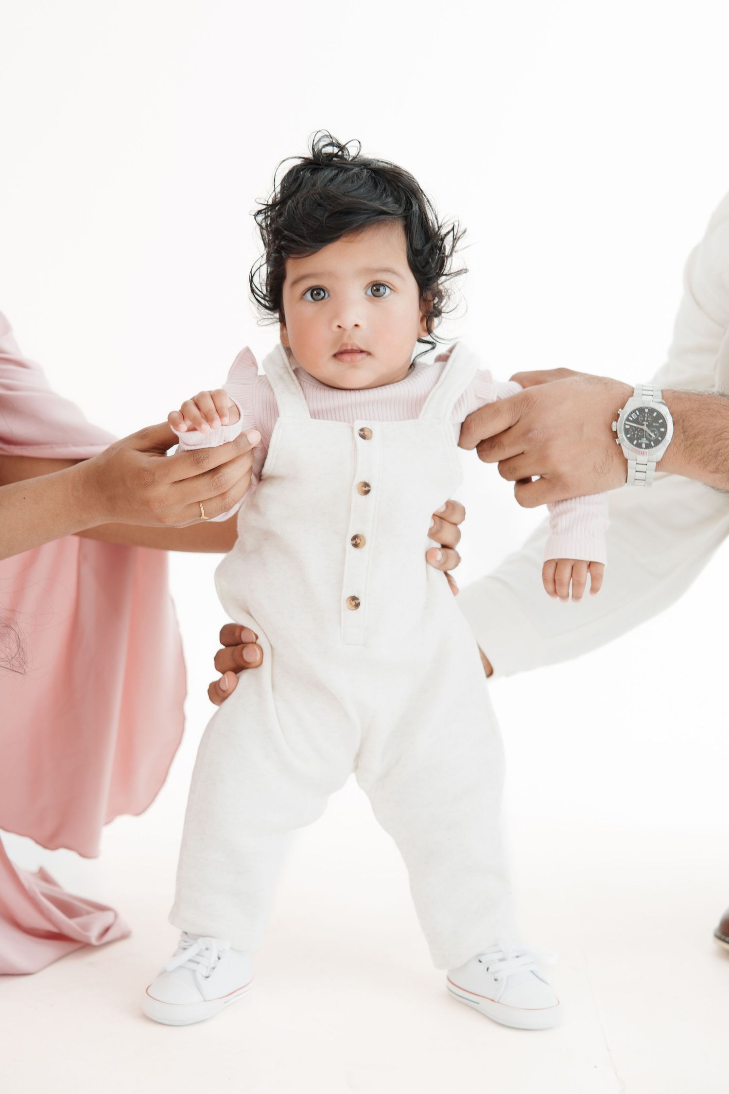 A baby in white outfit stands supported by parents hands on white background.