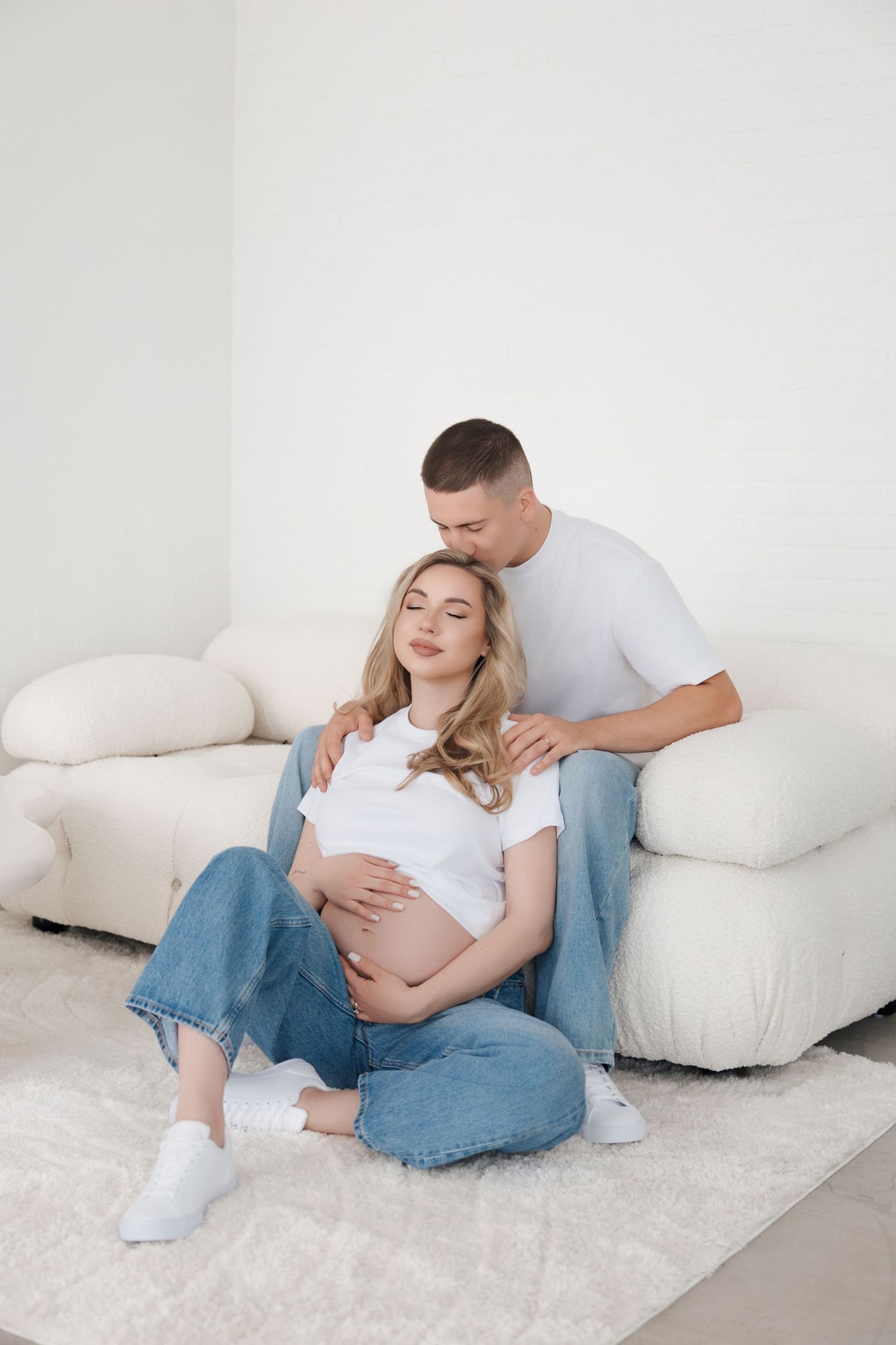 Pregnant woman sitting on floor leaning against couch while partner gives shoulder massage in bright white room.