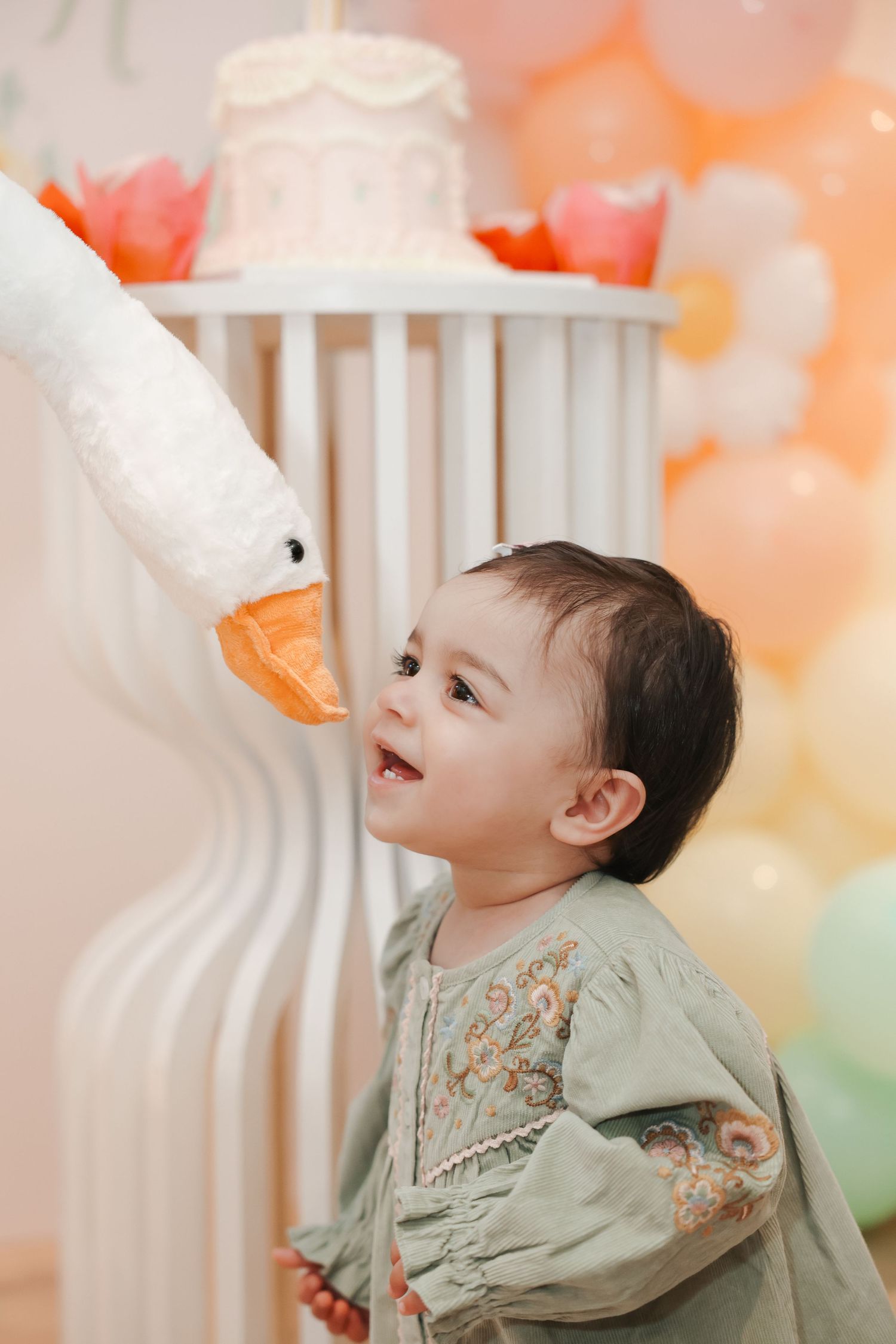 Toddler in green dress looks up at stuffed goose toy near birthday cake with pastel balloons in background.