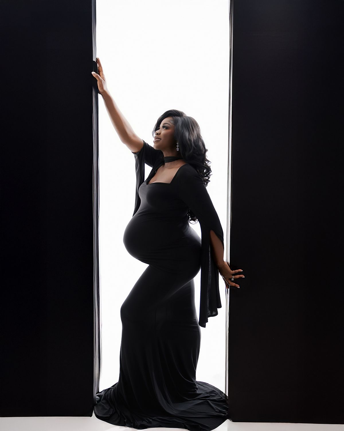 Pregnant woman in elegant black maternity gown posing dramatically in a bright doorway during a studio photoshoot.