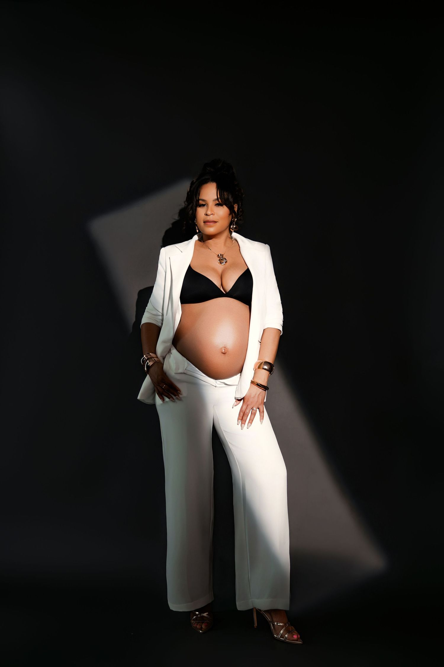 Pregnant woman in white suit jacket and pants with black bra, showcasing baby bump in dramatic studio lighting.