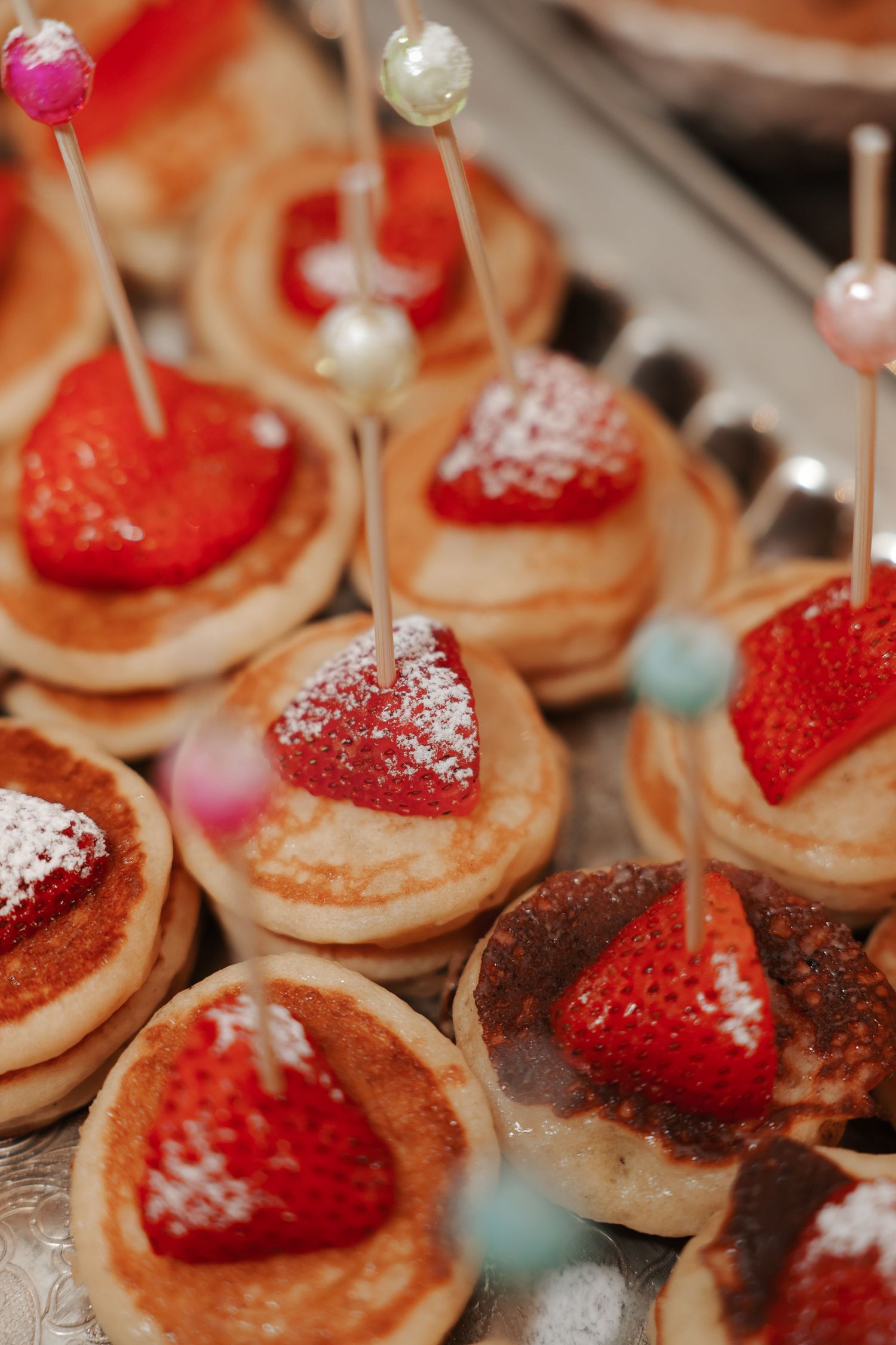 Mini pancakes topped with fresh strawberries and powdered sugar, garnished with decorative pearl picks on a silver tray.
