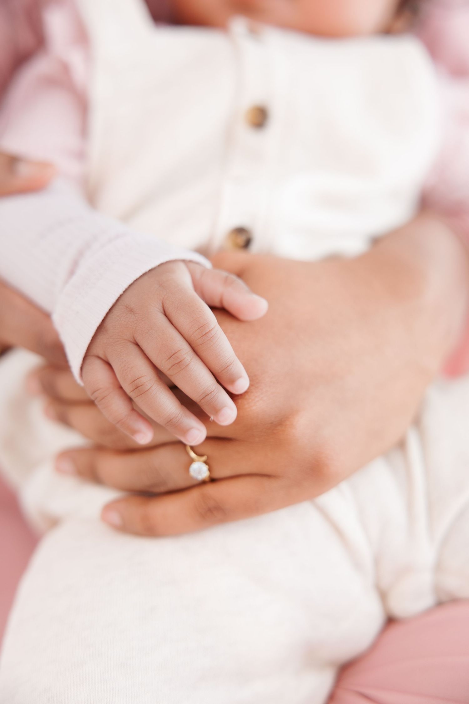A newborn baby's tiny hand resting on a parent's hand wearing a pearl ring, dressed in white.