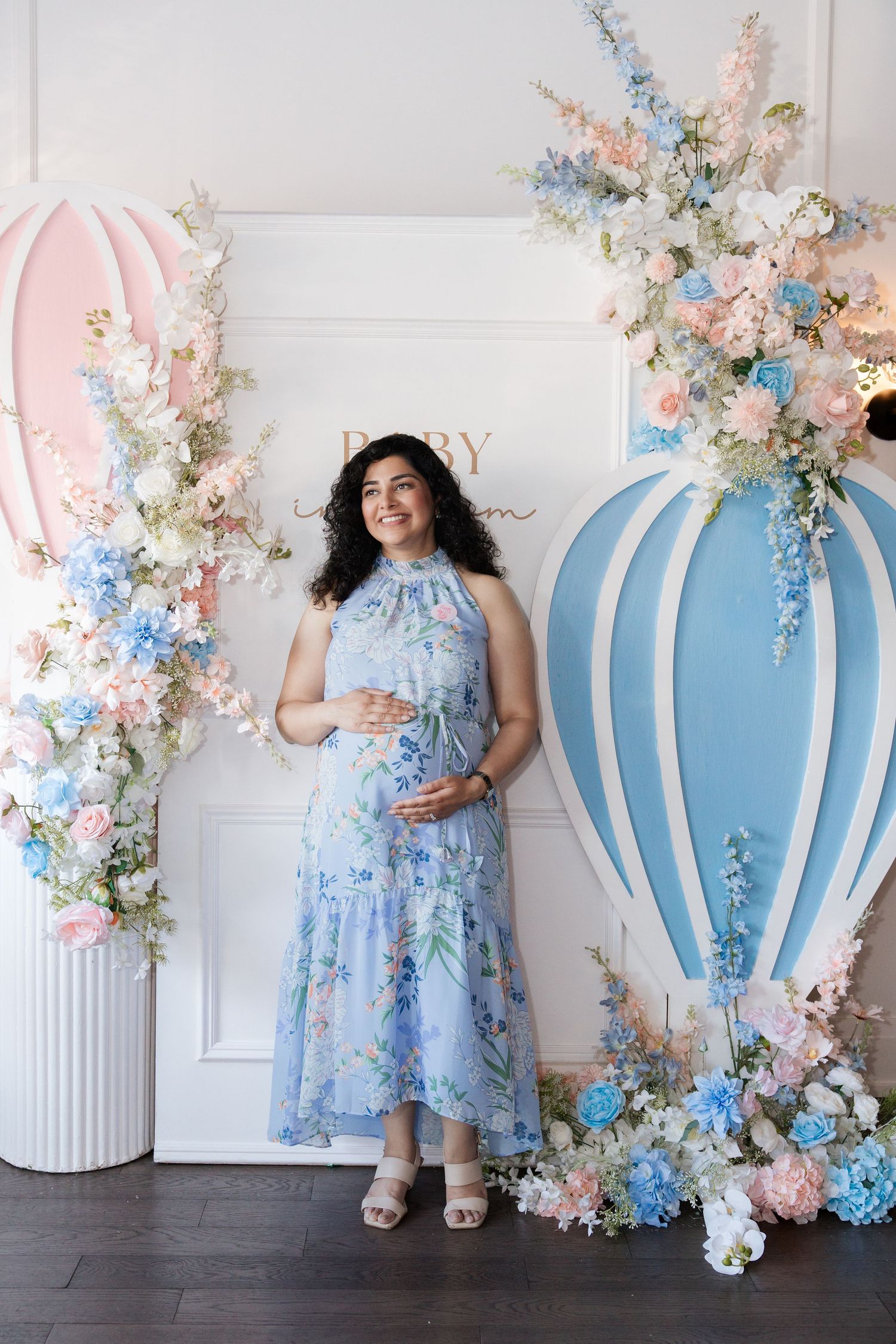 Smiling pregnant woman in blue floral dress poses at elegant baby shower with hot air balloon decor and flowers.