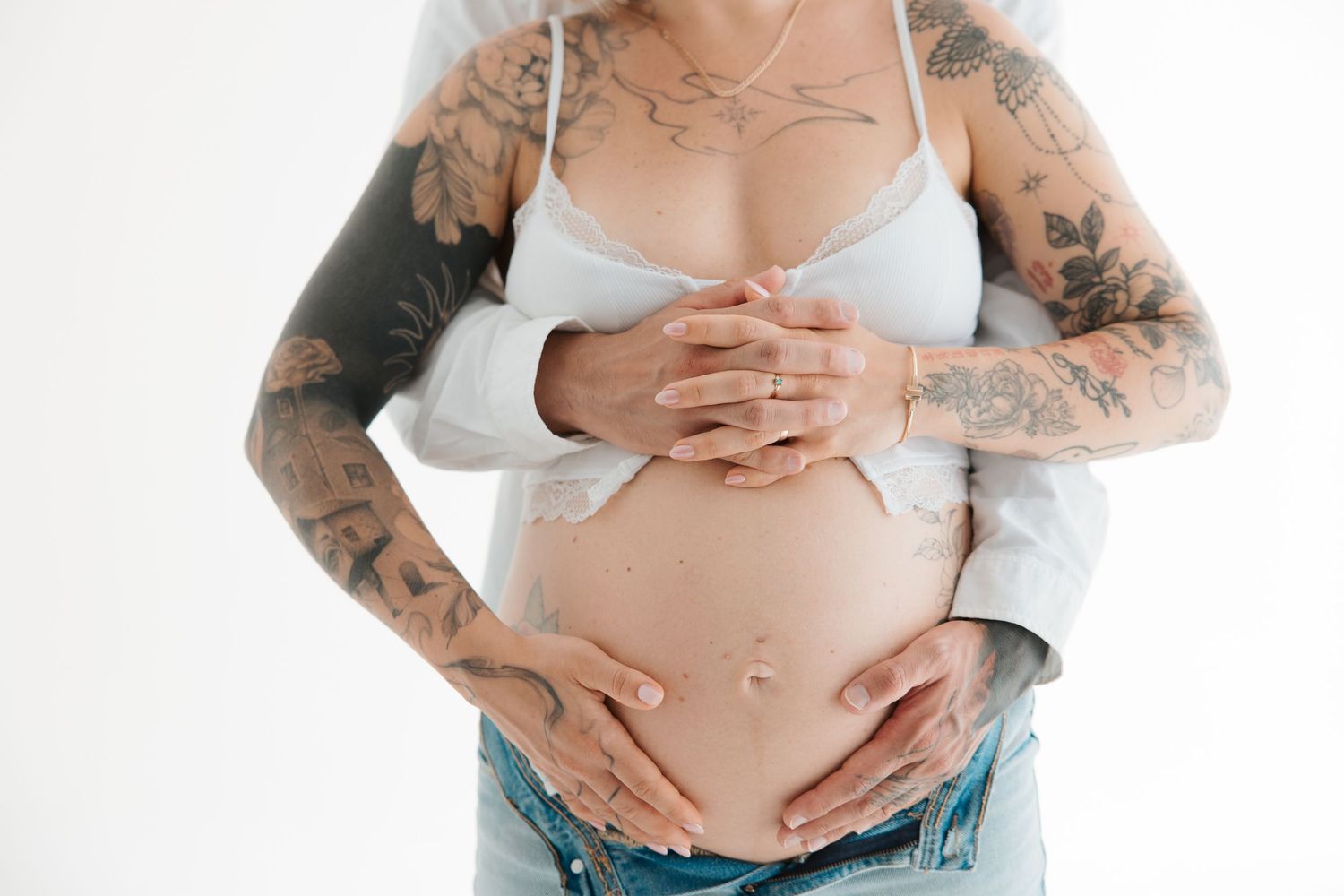 Pregnant woman with tattoos cradling baby bump, wearing white bra and jeans, partner embracing from behind.