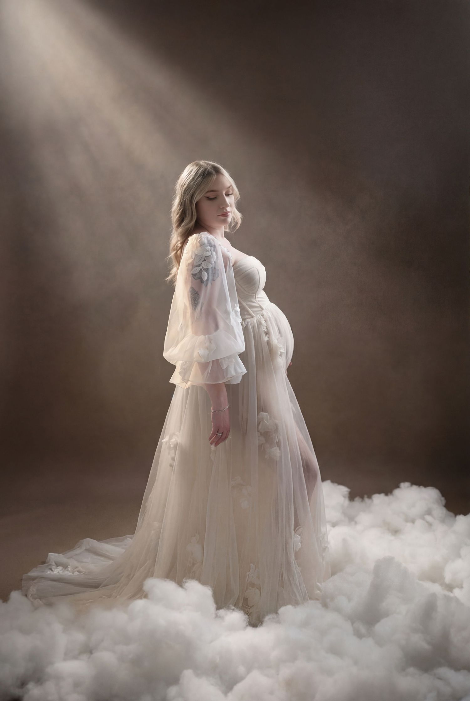 Pregnant woman in elegant white tulle gown with sheer sleeves, standing in dramatic light beam surrounded by clouds.