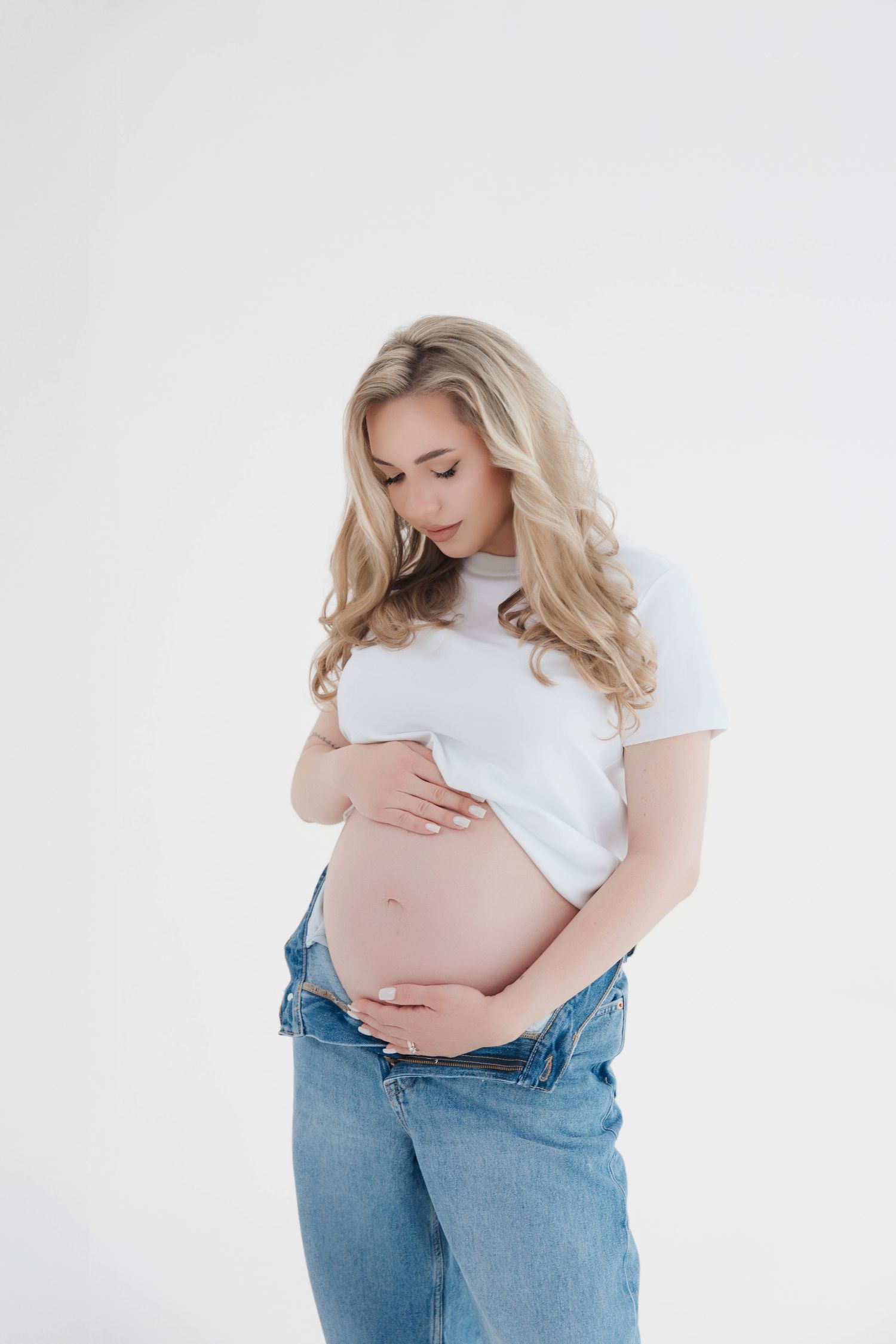 Pregnant woman in white t-shirt and jeans cradling her bare baby bump against a white background.