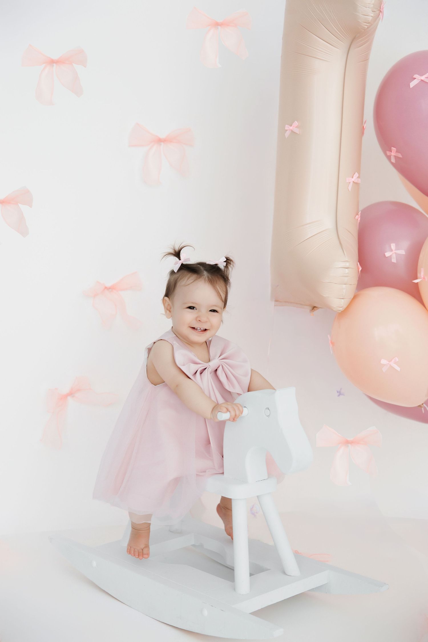 Smiling toddler girl in pink dress on white rocking horse, first birthday party with bow-decorated walls and balloons.