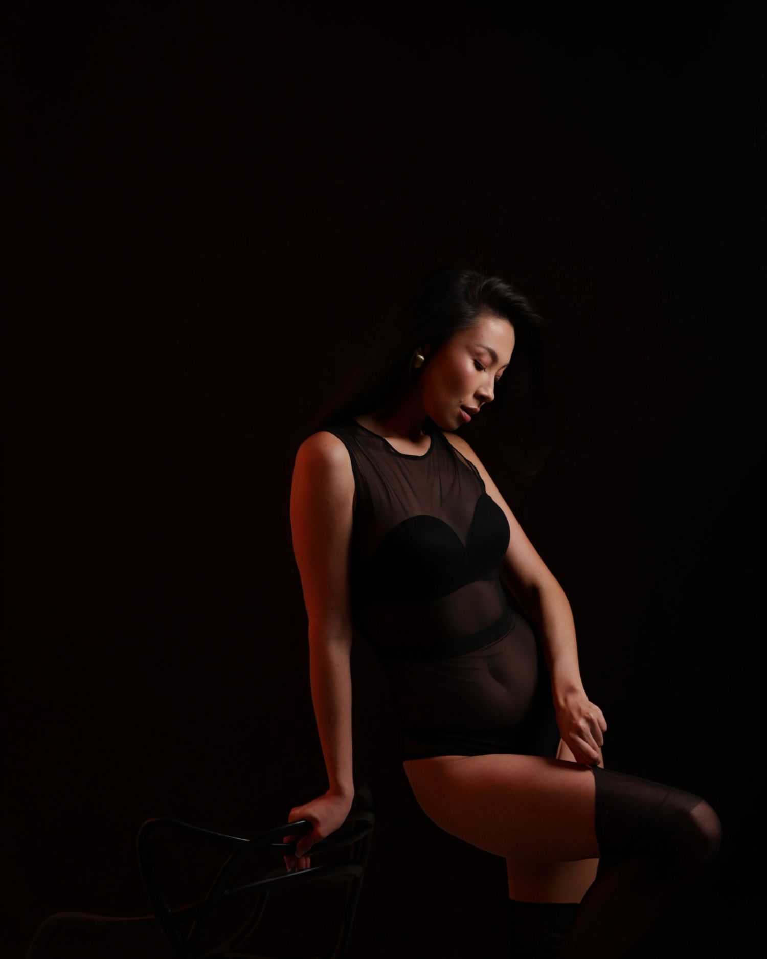 Woman in a sheer black bodysuit and stockings posing dramatically in low-key dark studio lighting.
