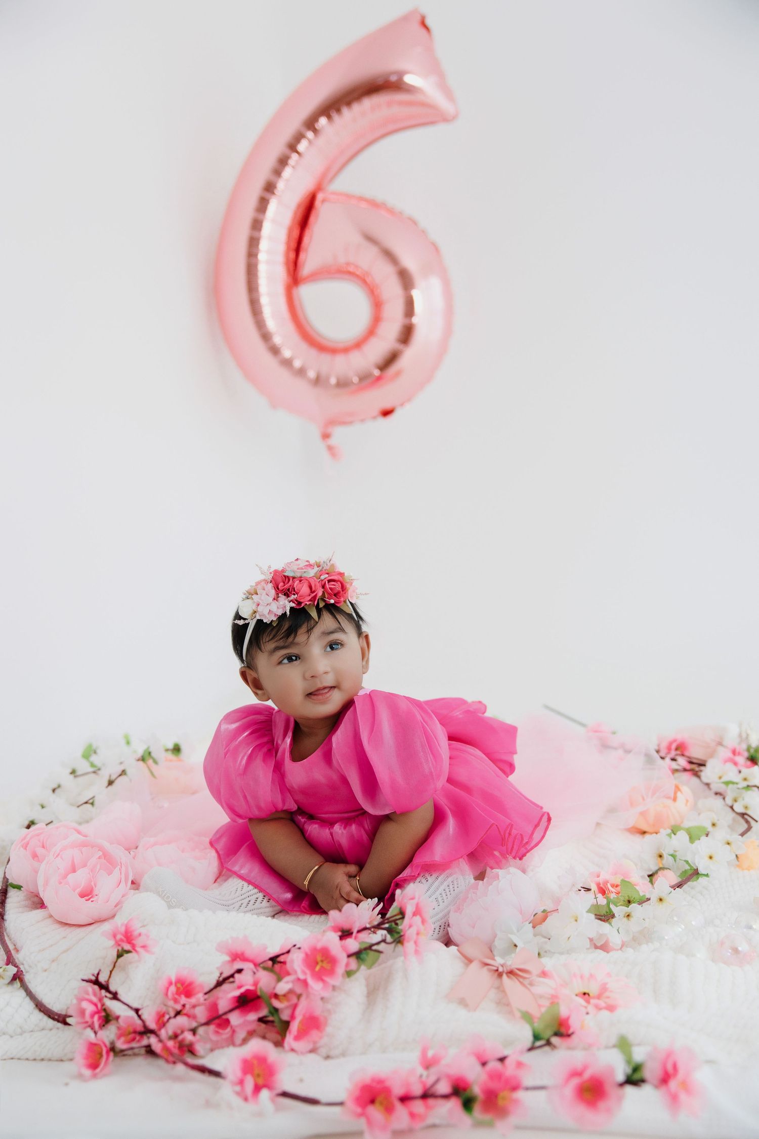 Baby girl in pink dress with floral headband sits among pink flowers under a number 6 balloon.