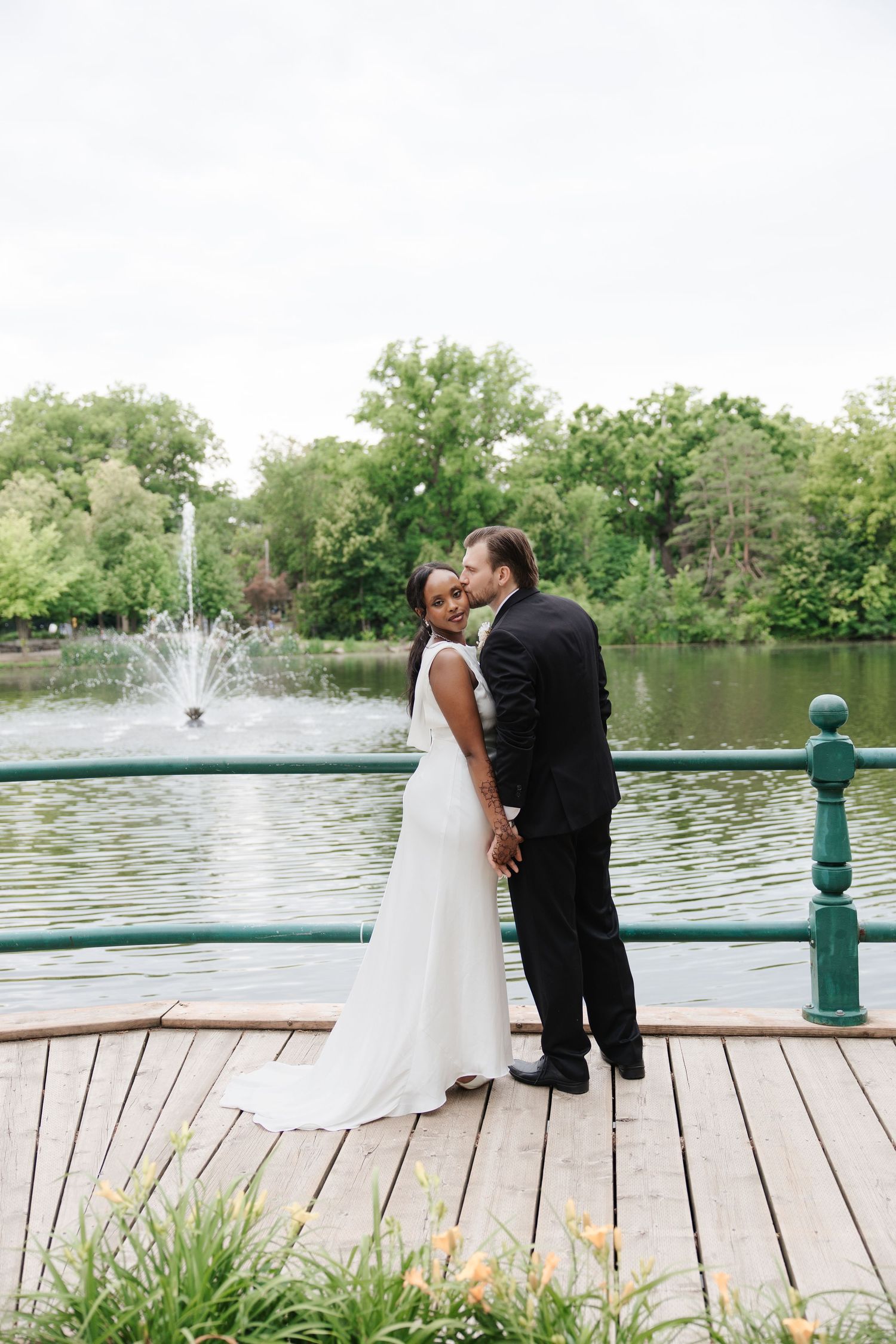 Interracial wedding couple posing on wooden dock by fountain and lake, bride in white gown, groom in black suit.