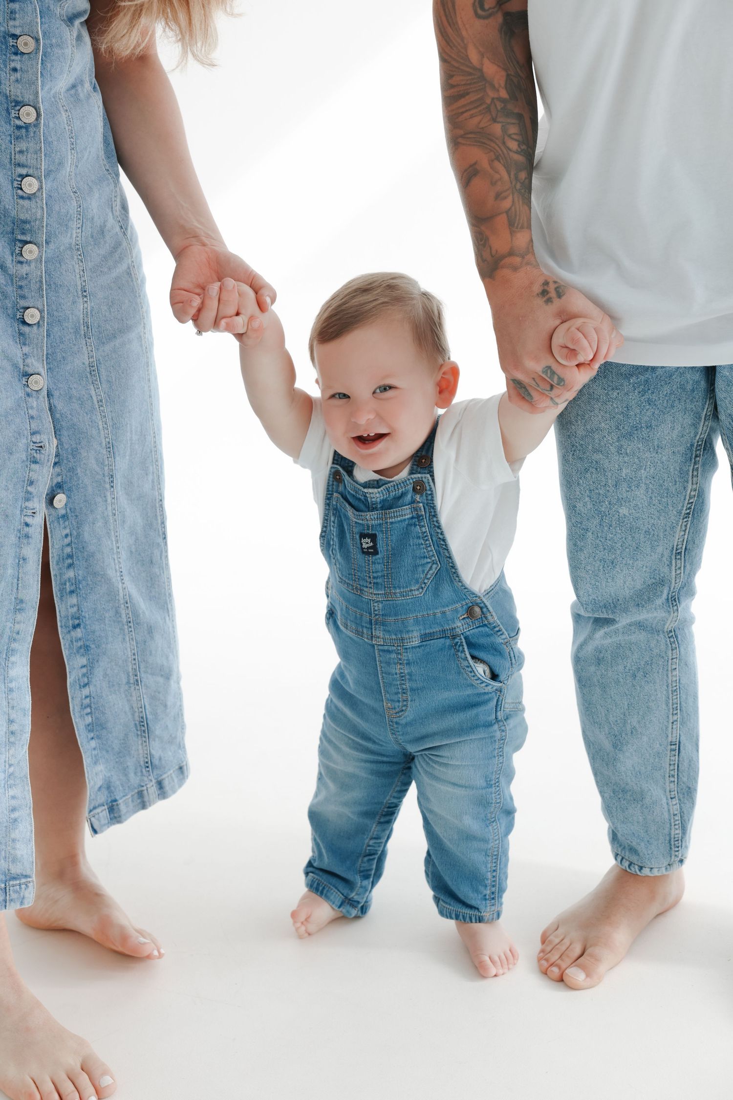 Happy baby in denim overalls takes first steps while holding hands with parents in matching denim outfits on white background.