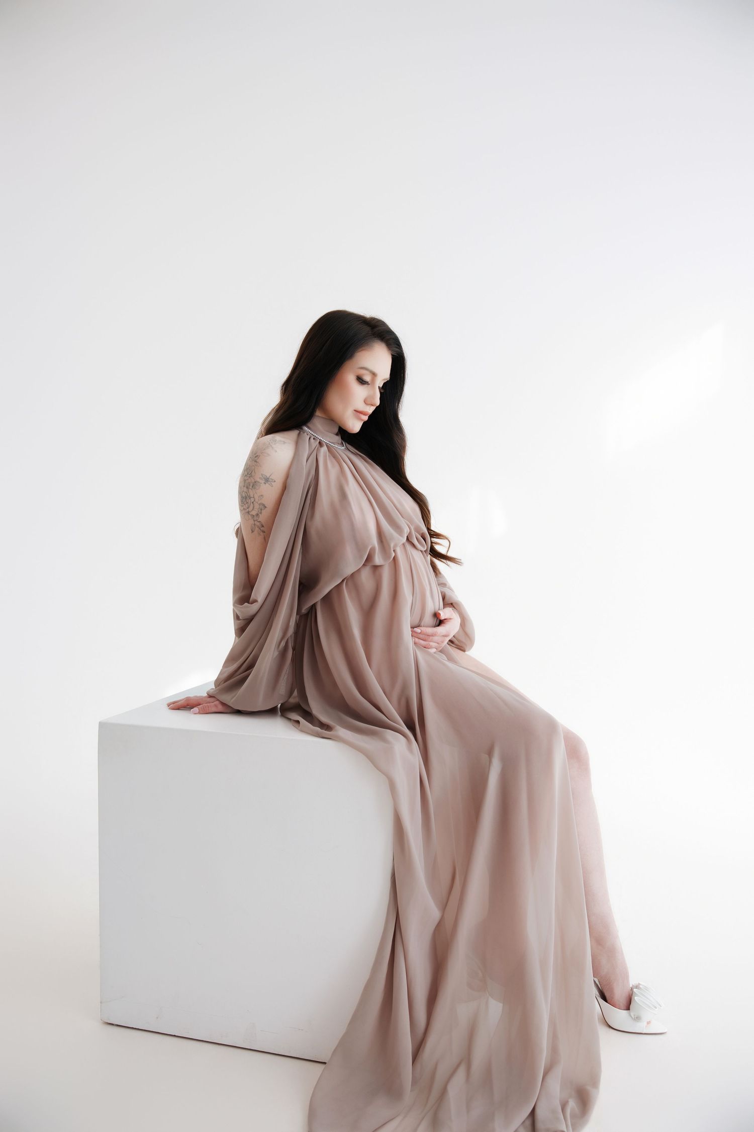 Pregnant woman in flowing beige dress sitting gracefully on white cube, cradling baby bump in bright studio setting.