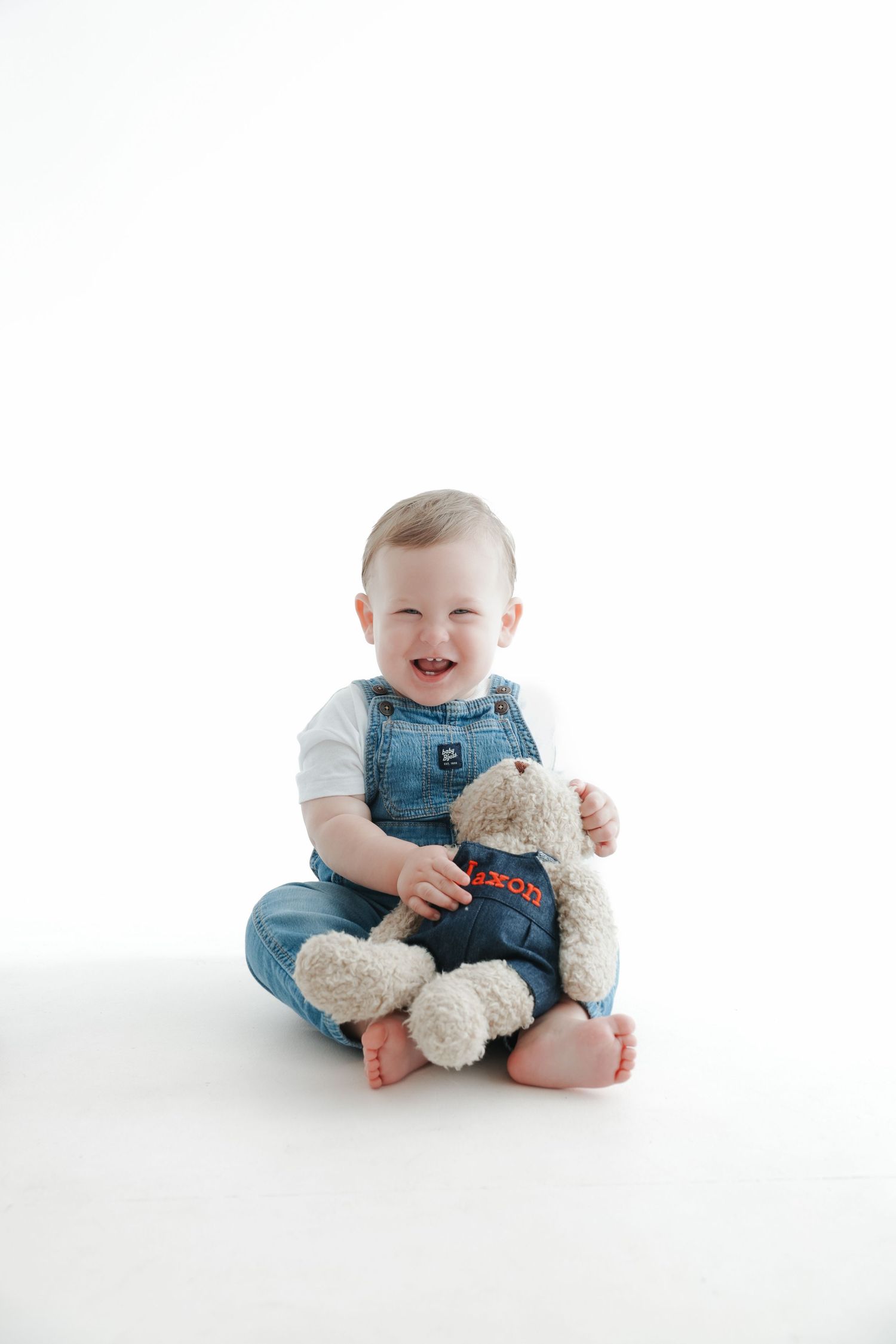 Smiling baby in denim overalls sitting on white background holding a personalized teddy bear.